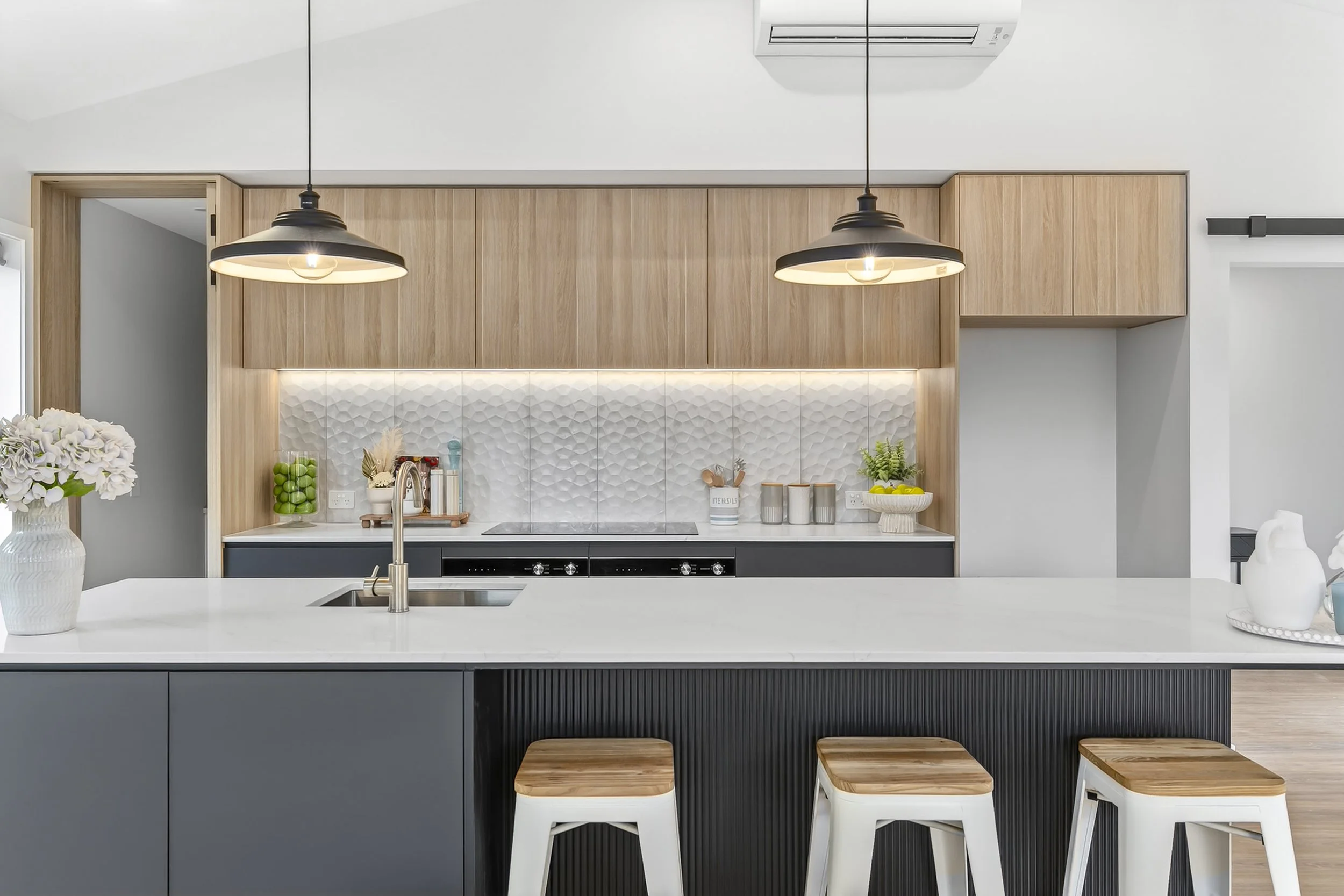 Modern kitchen with a white island counter, three white stools with wooden seats, wooden cabinets, white textured backsplash, and black pendant lights hanging above.