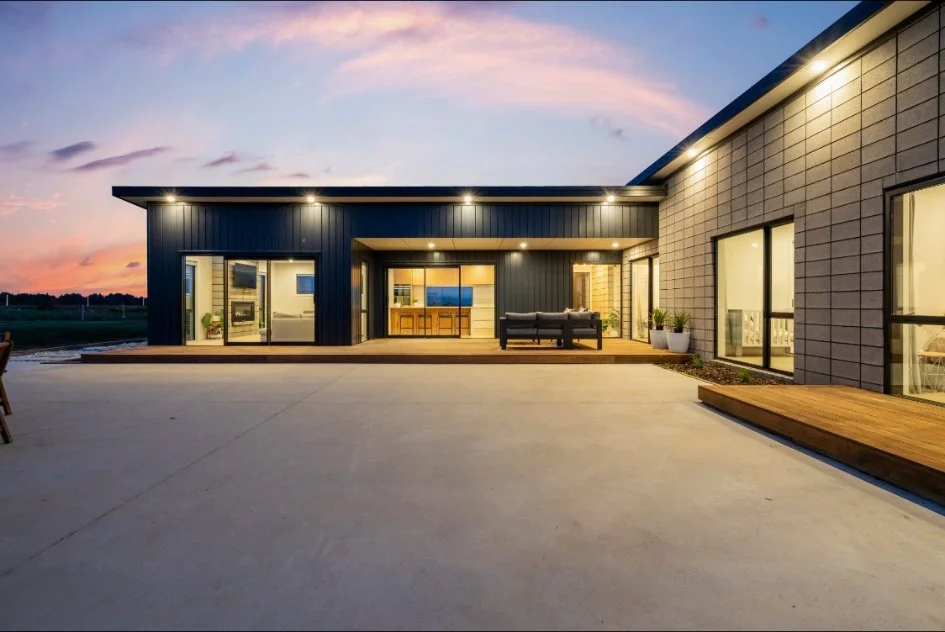 Modern house exterior during sunset with large patio area, sliding glass doors, and outdoor seating.