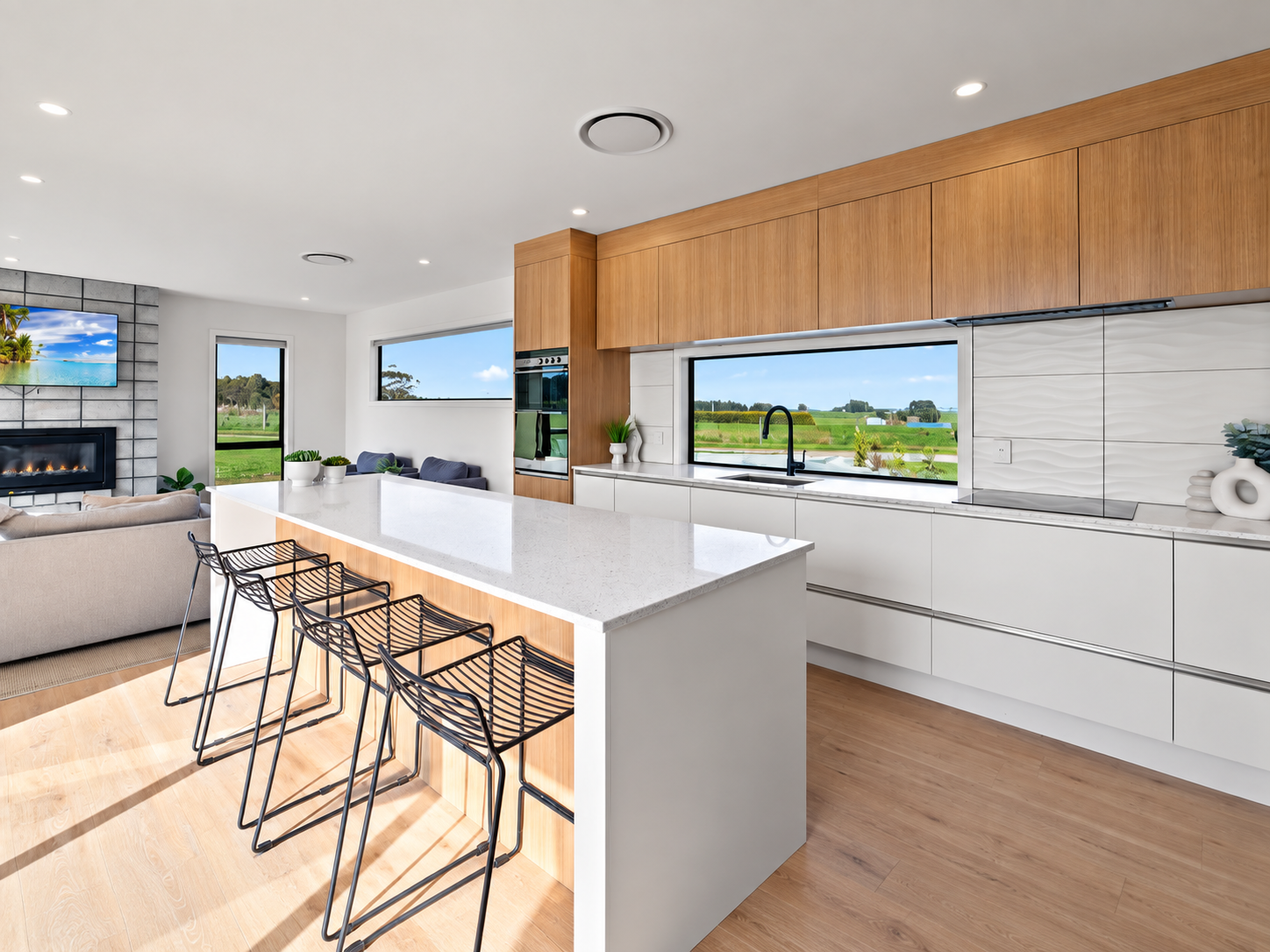 Modern kitchen with white countertops, wooden cabinets, black faucet, and large window showing green fields.