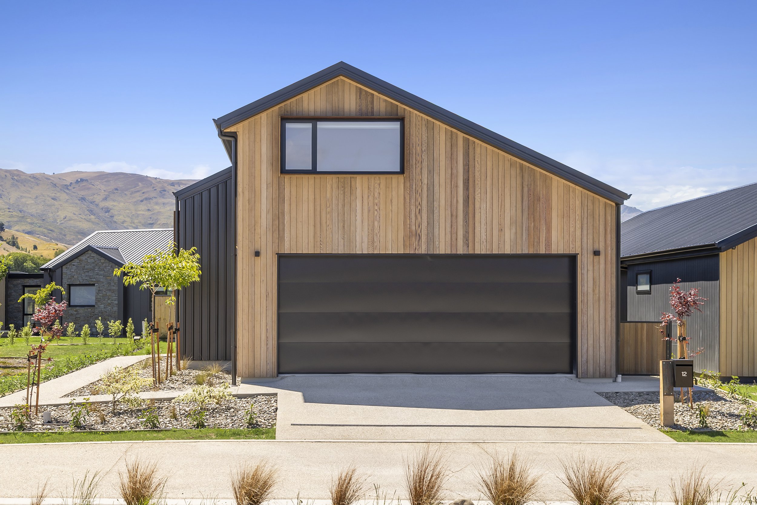 Modern two-story house with a wood and black exterior, large window near the roof, driveway, and landscaped yard with small trees and plants, set against mountains and a bright blue sky.