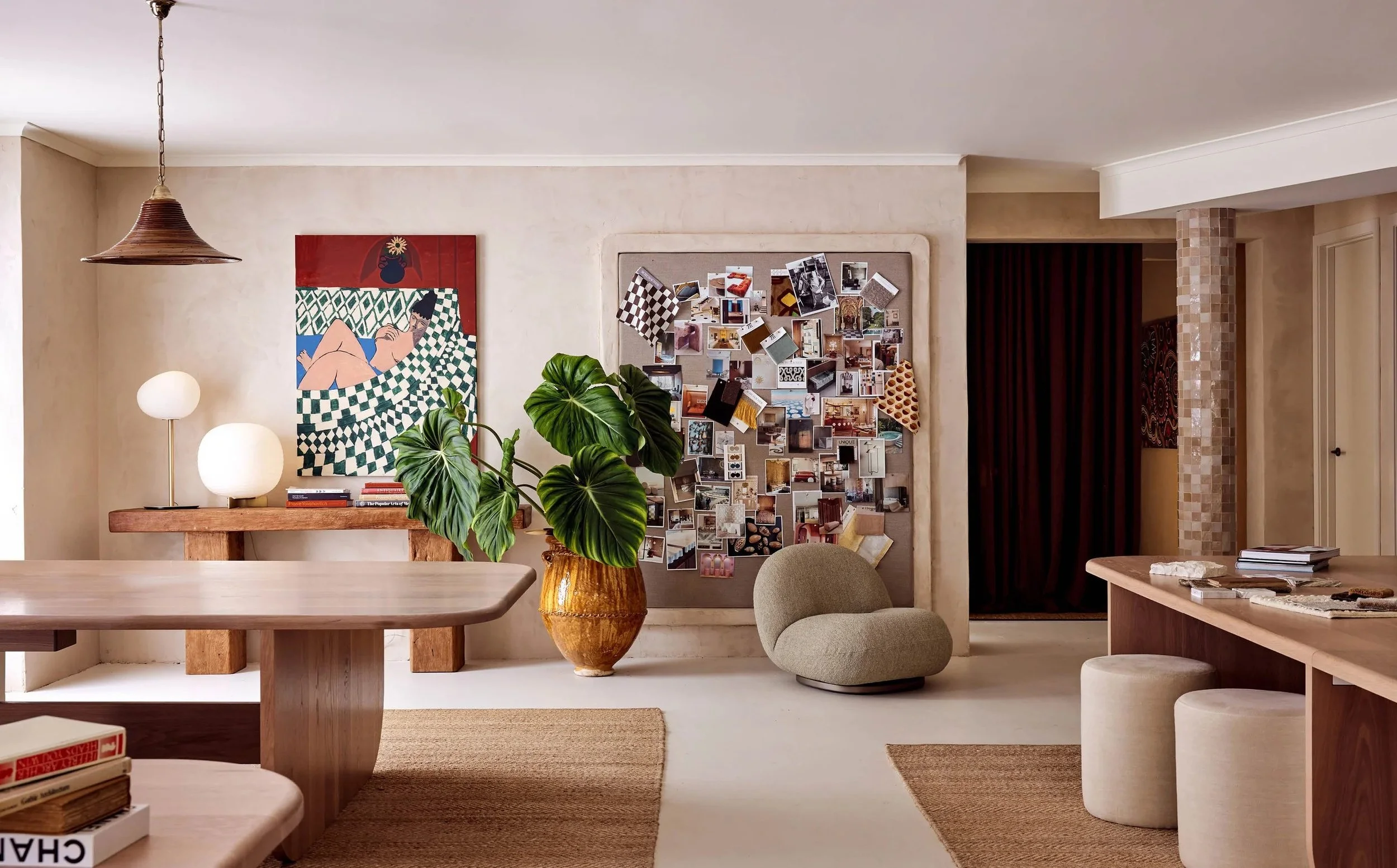 Living room with beige walls, wooden furniture, a large potted plant, wall art, a privacy board with photos, a cushioned chair, and small stools.
