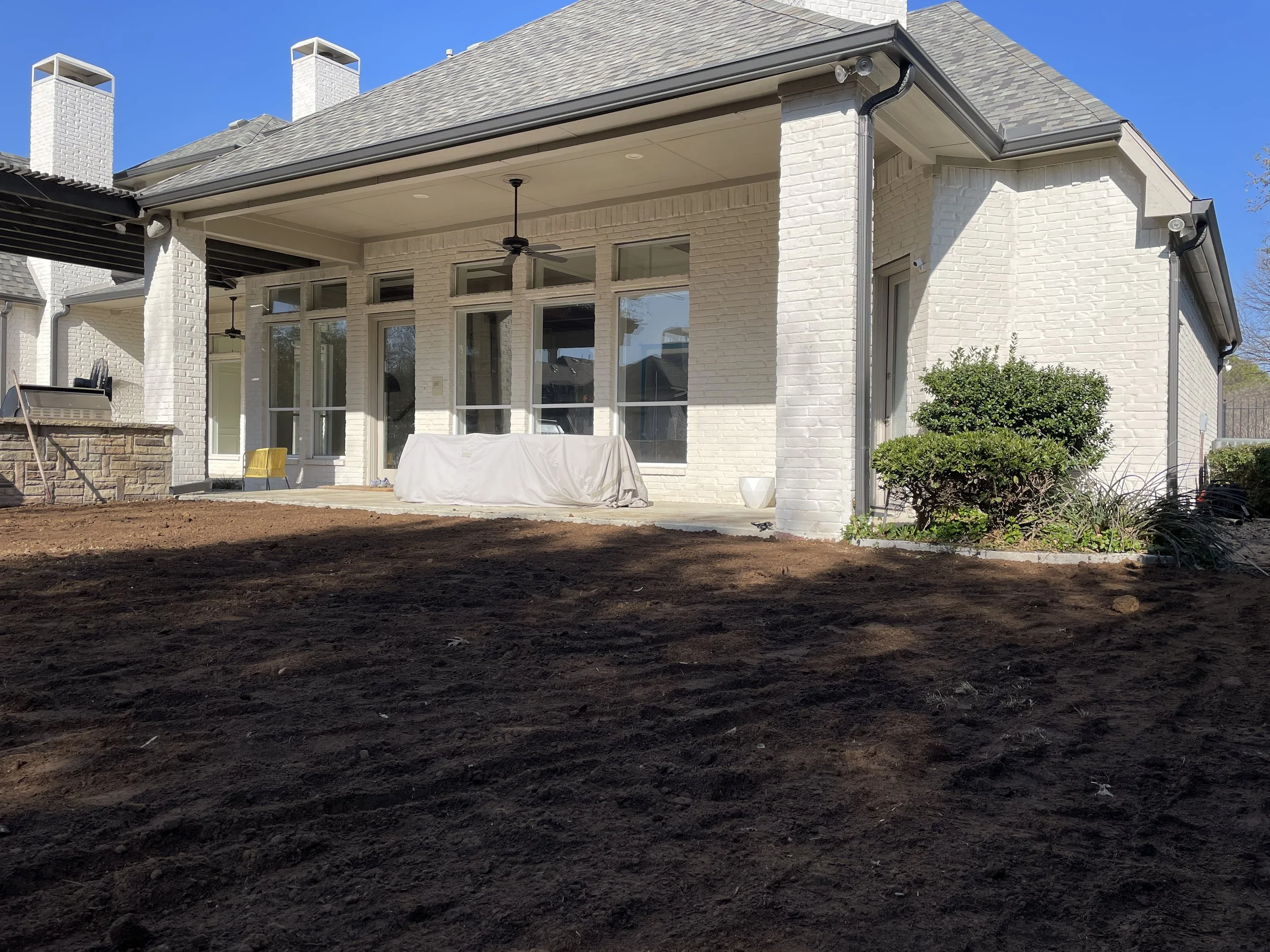 Backyard of a white brick house with a covered patio, ceiling fans, and large windows, showing a freshly tilled soil yard and some landscaping bushes.
