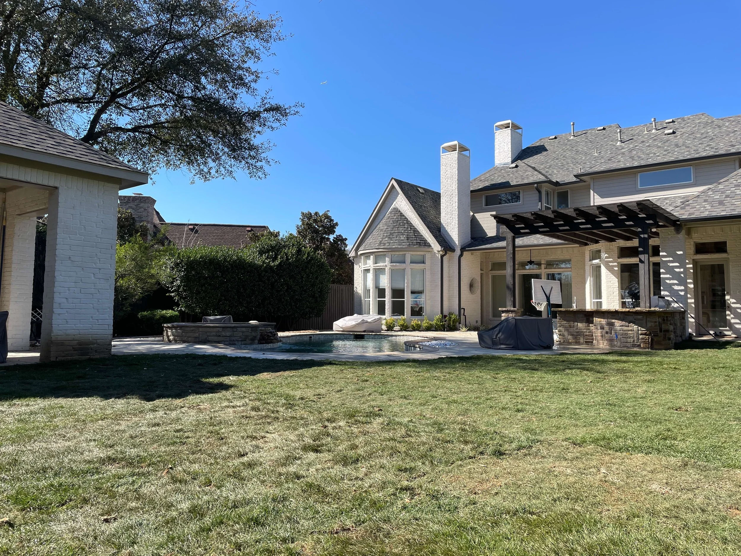 Backyard view of a large house with a patio, swimming pool, and a grassy lawn under a clear blue sky.