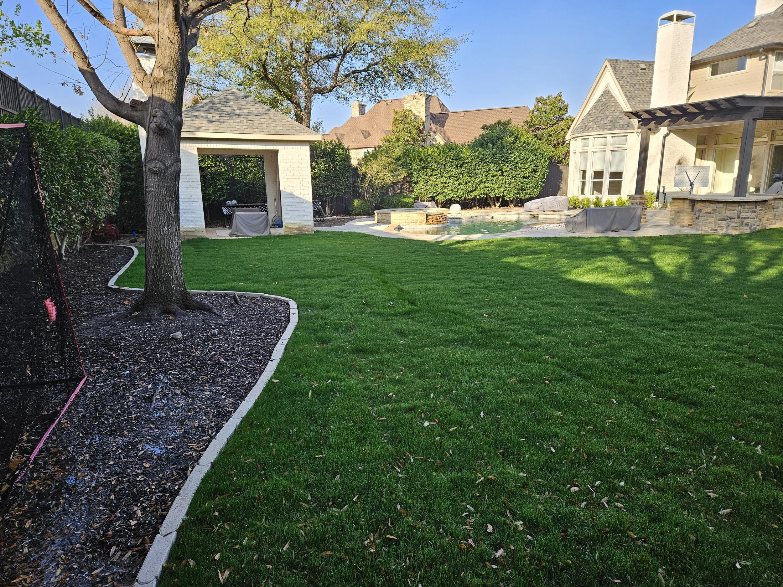 Backyard with neatly mowed grass, trees, a small pool, and a house with a white exterior and stone accents.