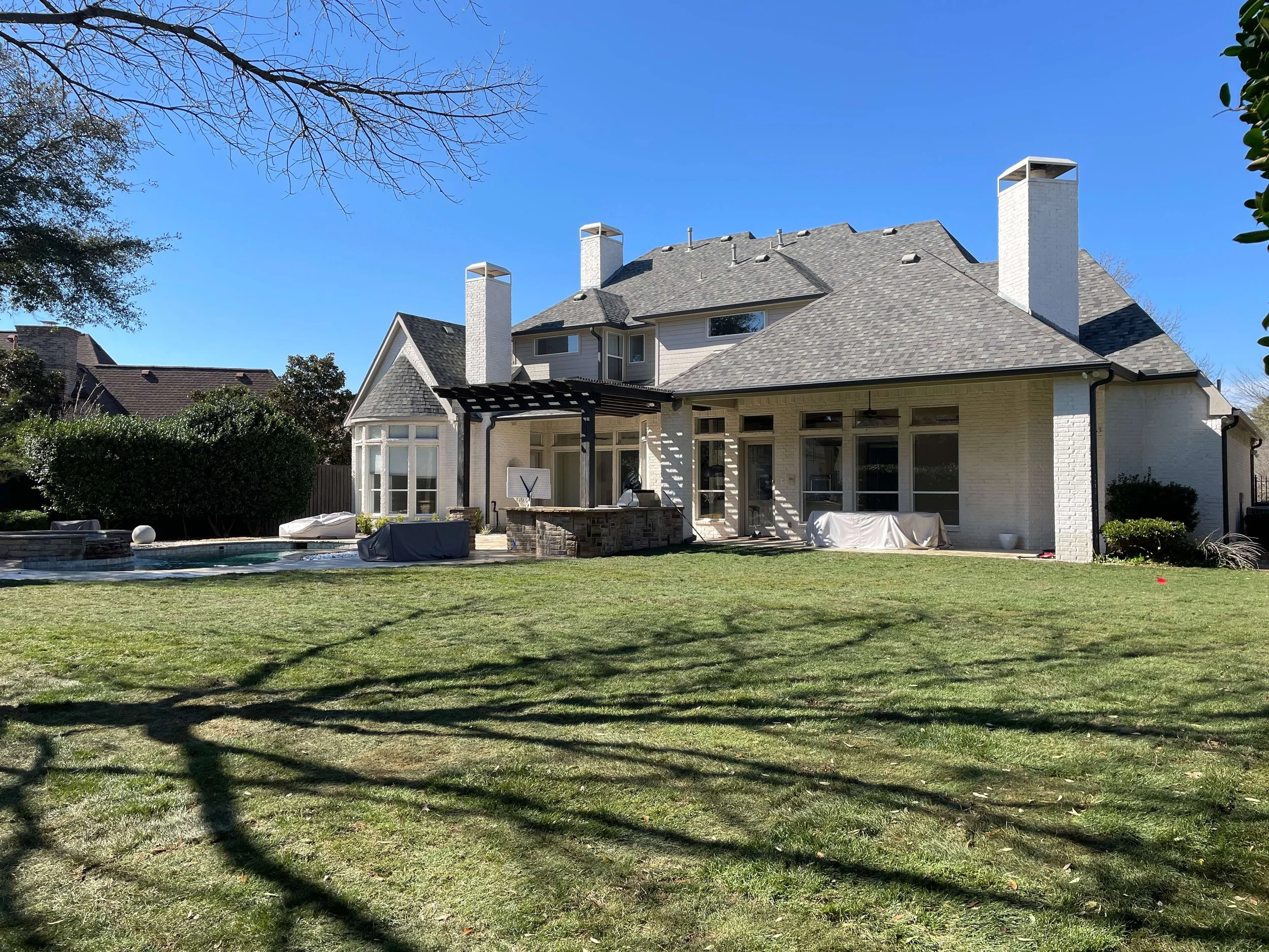 Backyard view of a large white house with multiple chimneys, a gray shingled roof, and large windows. There is a green lawn, a patio with outdoor furniture, and some covered items. Trees and neighboring houses are visible in the background.