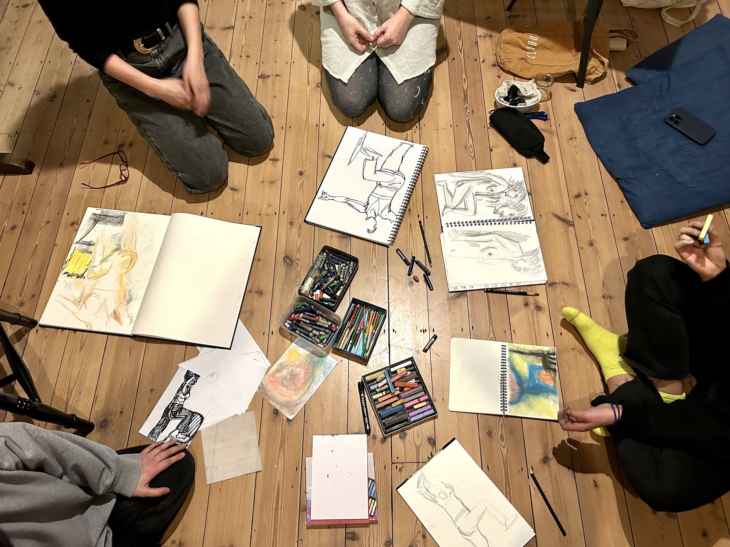 People sitting on a wooden floor with sketchbooks, drawing supplies, and artwork, engaged in an art session or class.