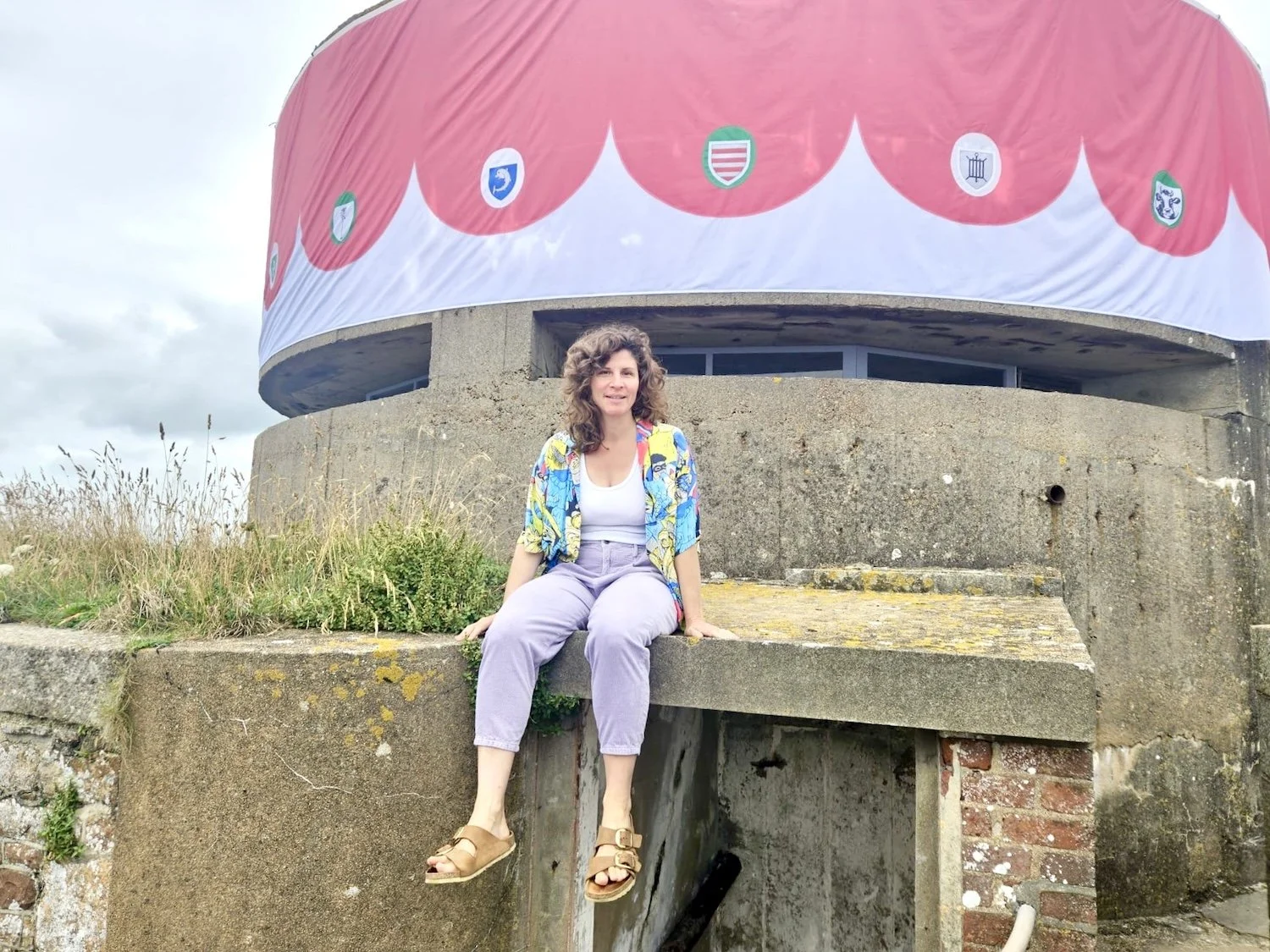 Woman sitting on concrete ledge in front of a round building with a red and white striped awning featuring badges or coats of arms.