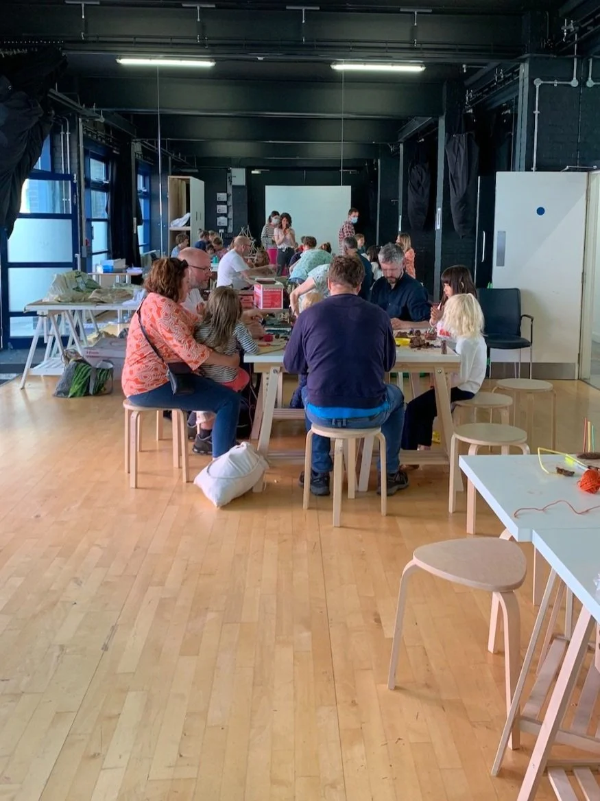 People participating in a group activity or workshop at tables in a well-lit indoor space with wooden floors, black walls, and large windows.