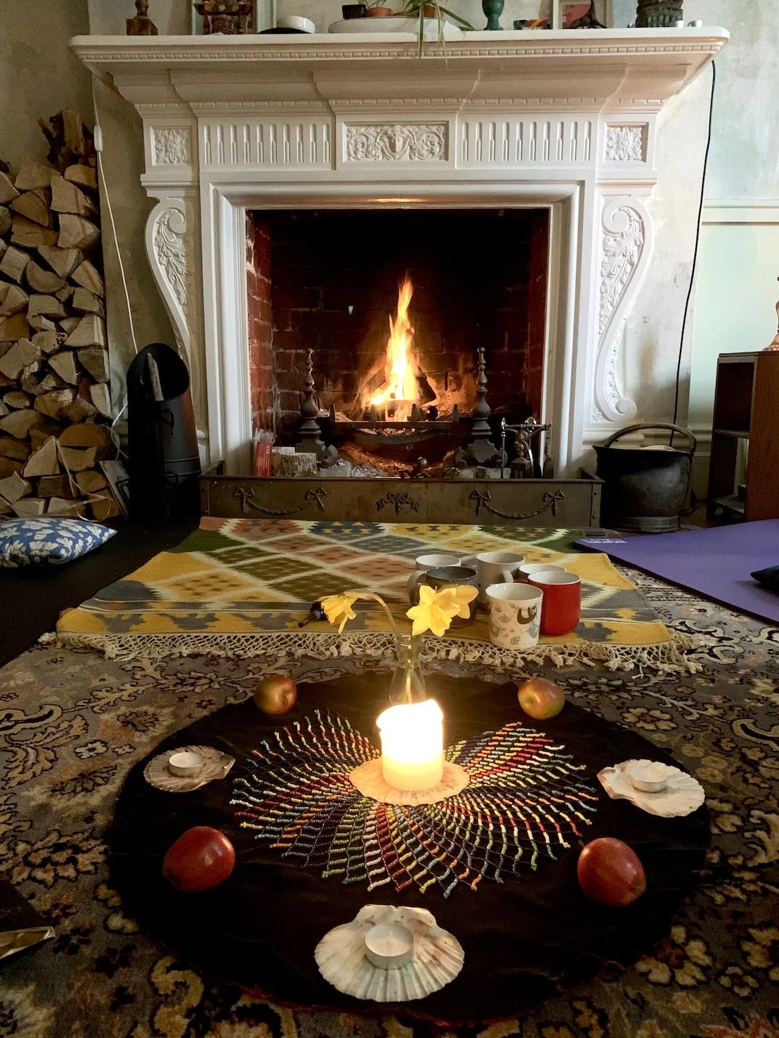 A cozy living room with a lit fireplace, decorated with a rainbow-colored thread design on a black cloth, four apples, and four tealight candles. There are cups on a yellow and multicolored cloth, with a stack of firewood to the left and decorative items on the mantelpiece.