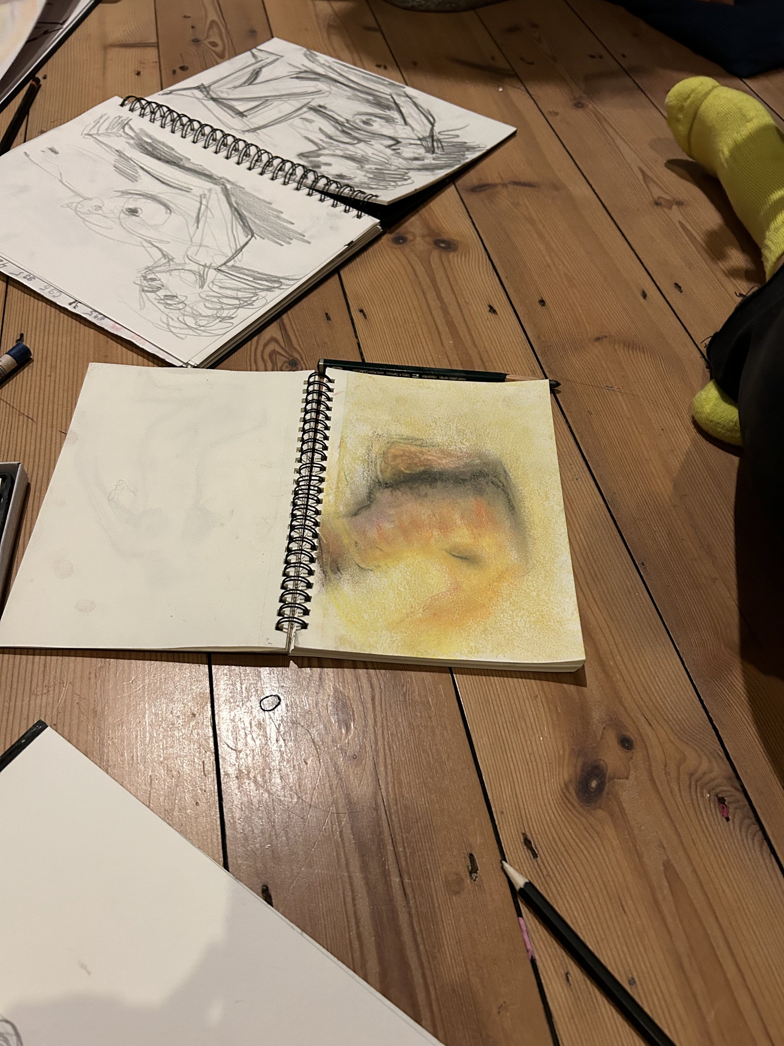 Open sketchbooks on a wooden floor, one with a detailed colored portrait and another with pencil sketches of a face, with various drawing tools nearby.