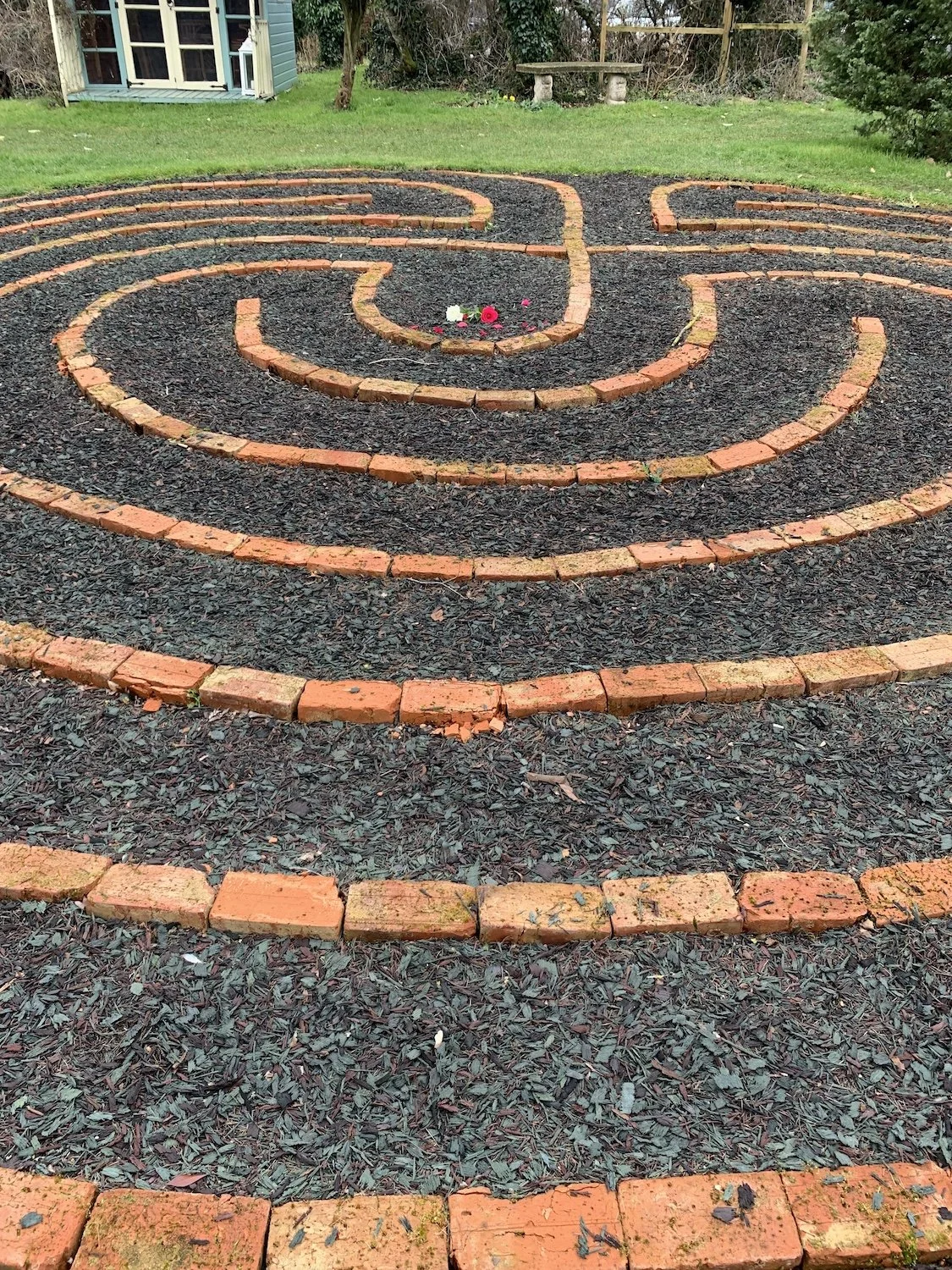 A backyard garden maze made of low brick walls with black gravel in between, featuring a spiral design with a small flower bed at the center. There is a green lawn, a small blue shed, and some patio benches in the background.