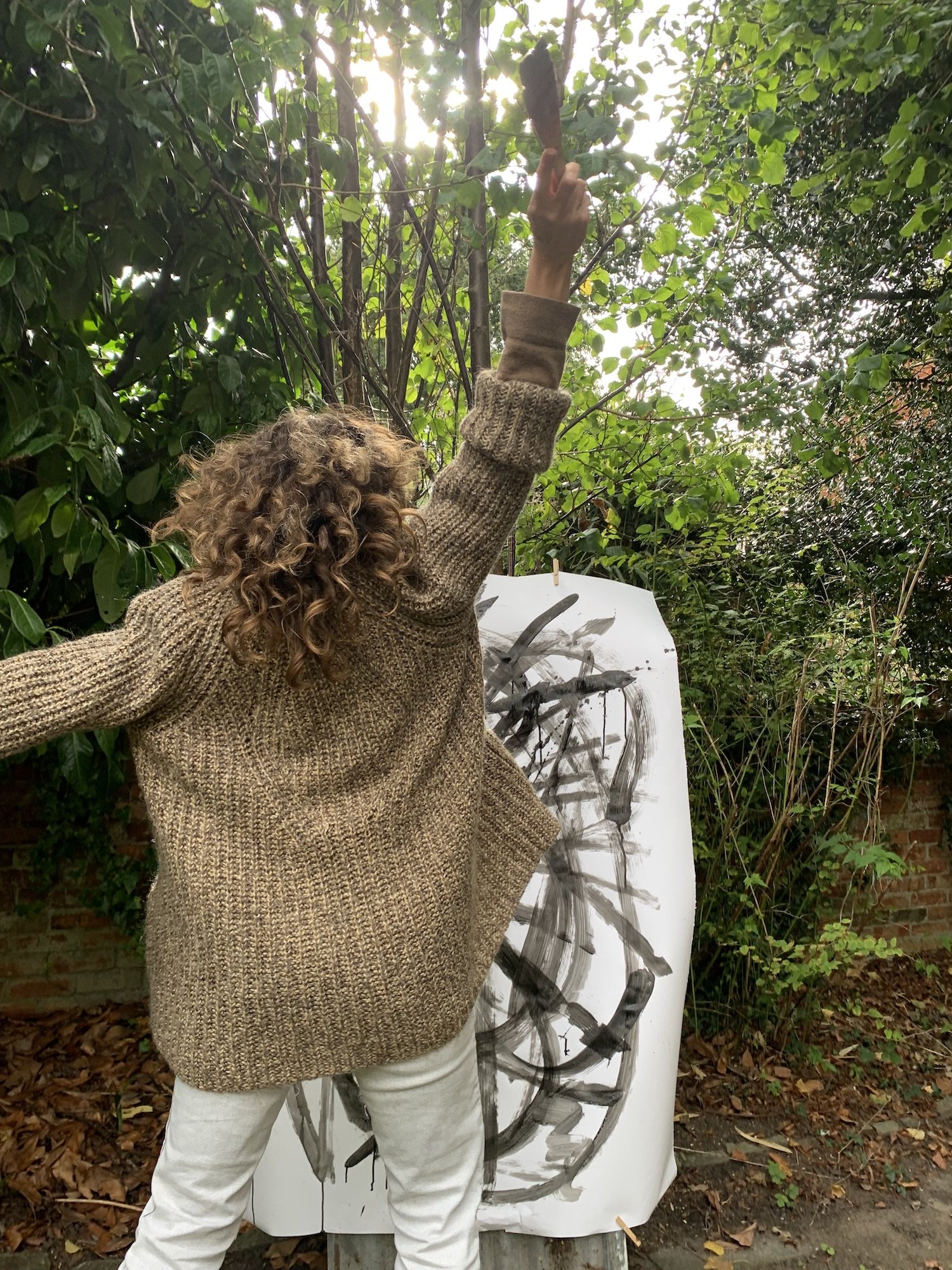 Person with curly hair wearing a brown sweater painting on a large white canvas outdoors surrounded by greenery.