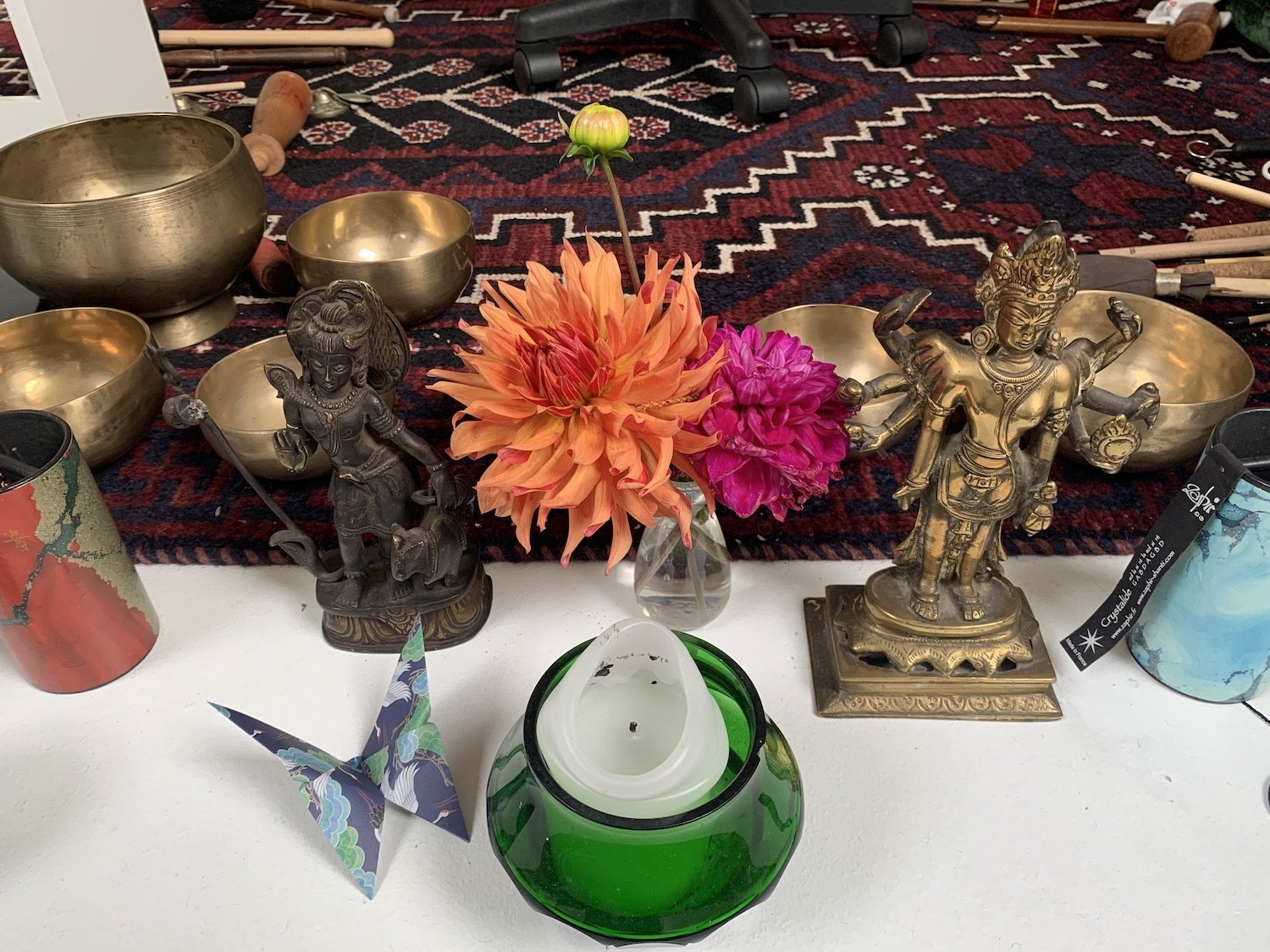 Arrangement of Hindu deities figurines, brass bowls, colorful flowers in a glass vase, and a green candle holder on a white surface with a patterned rug in the background.