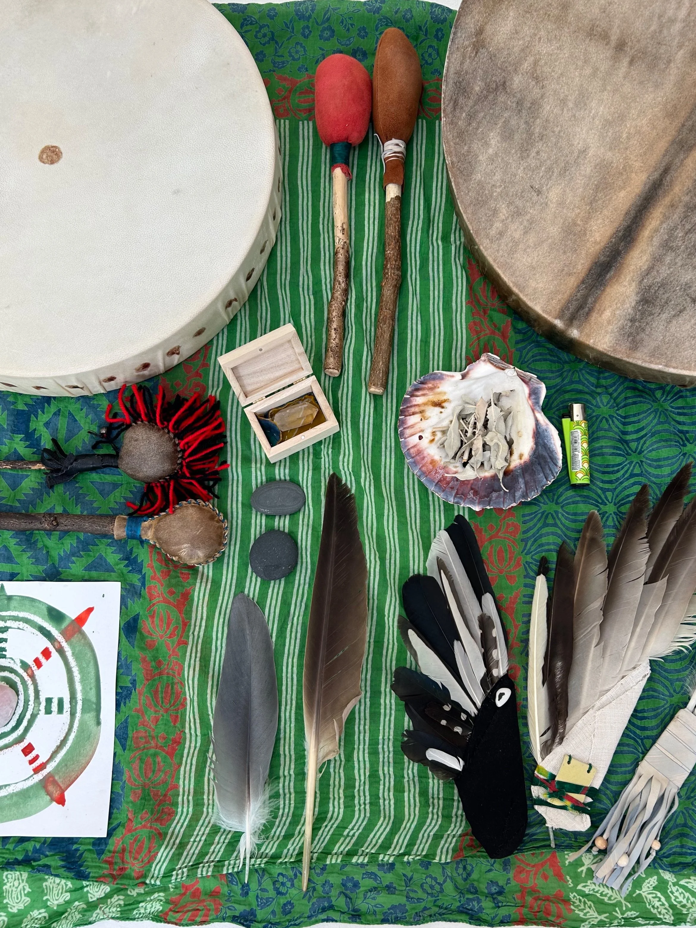 Display of Native American and Indigenous artifacts including feathers, drums, a shell, stones, and small tools on a green patterned cloth.