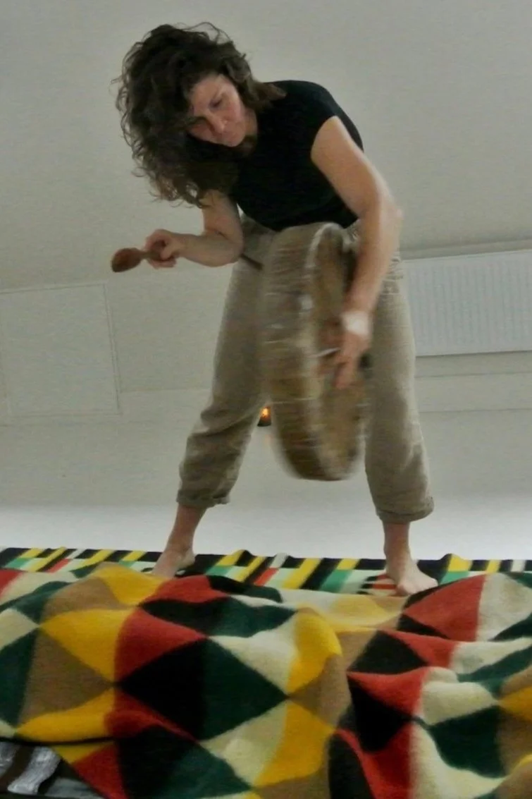 Woman with curly hair standing on a bed, holding a wooden spoon in one hand and a large wooden bowl in the other, surrounded by colorful blankets.