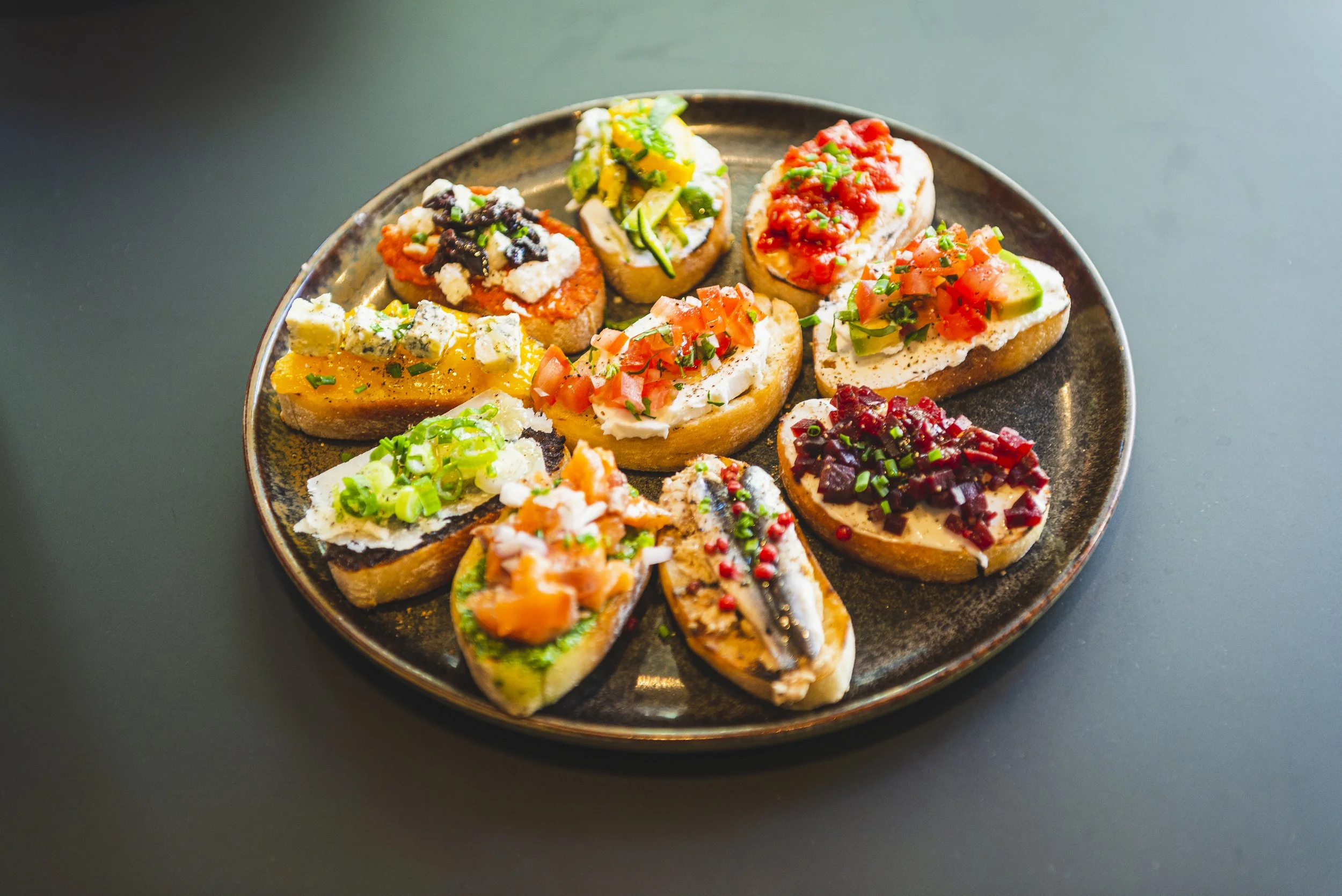 Een ronde black plate met verschillende soorten tapas met brood, zoals gerookte vis, paddestoelen, tomaat, komkommer, en kaas.
