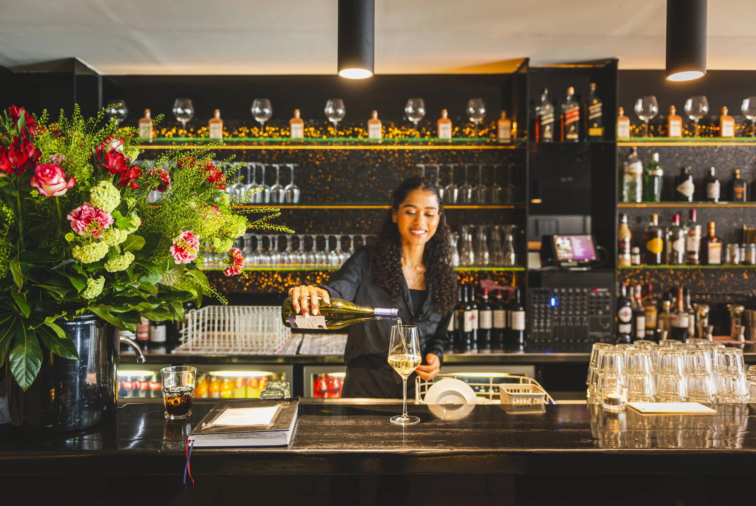 Vrouw die wijn schenkt in een bar met bloemen en glazen op de achtergrond.