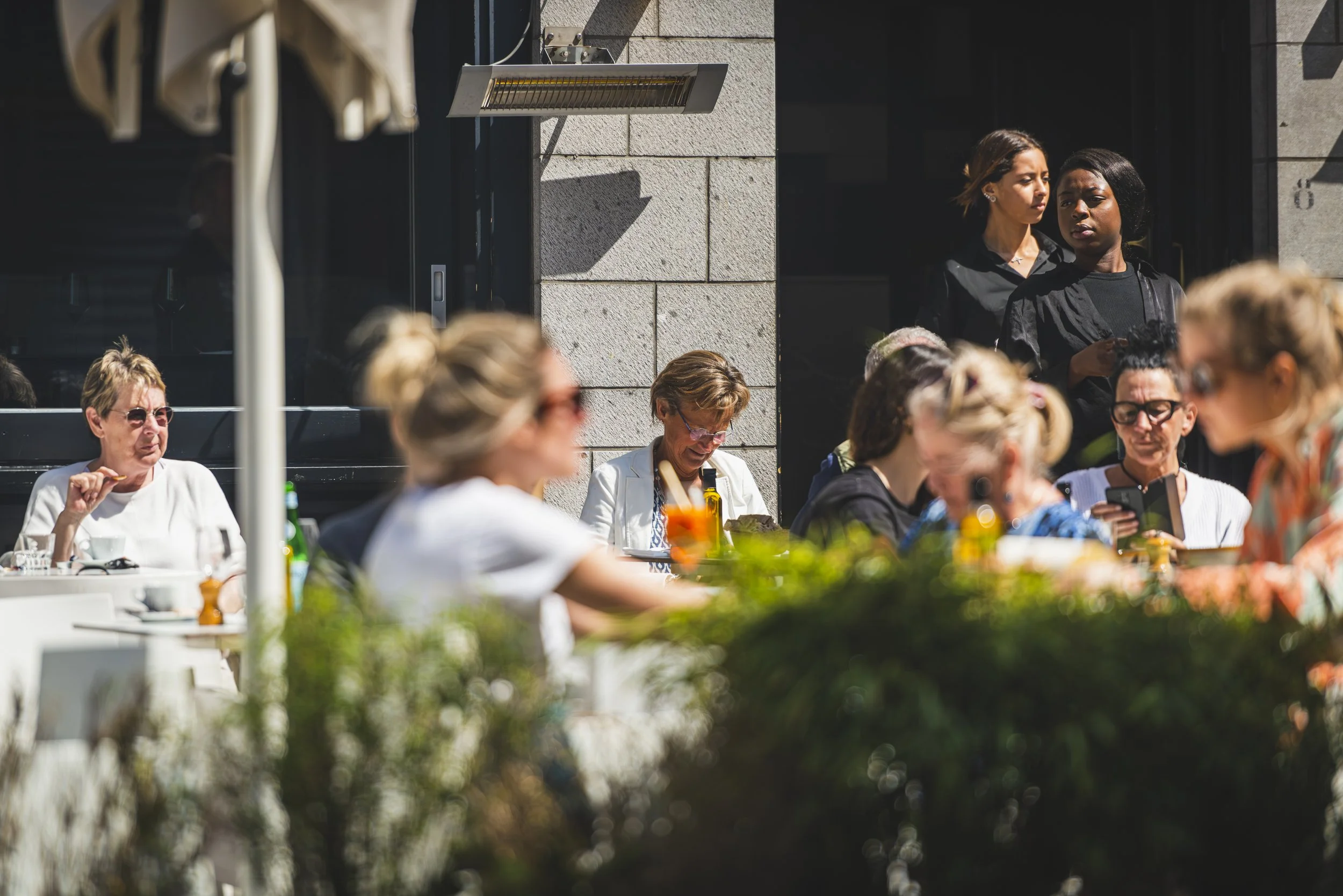 Mensen zitten en staan buiten op een zonnige dag voor een restaurant of café, met sommigen die hun telefoons gebruiken, terwijl anderen praten of genieten van het uitzicht.