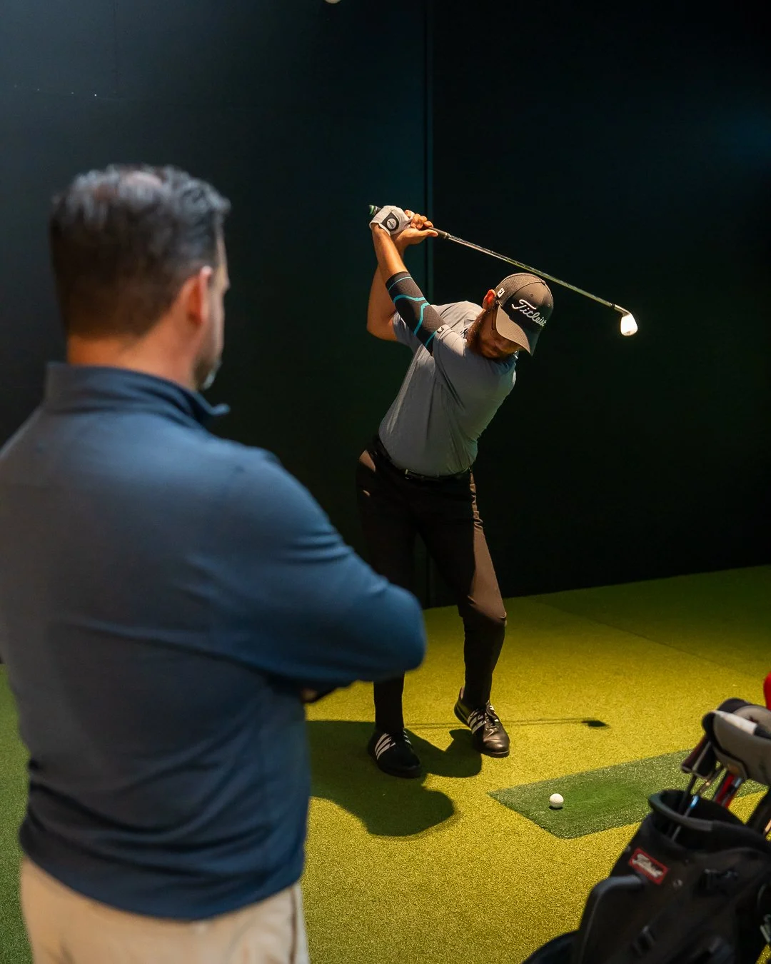 A man practicing golf swings in an indoor golf simulation room, while another man observes, with golf clubs and a golf bag nearby.