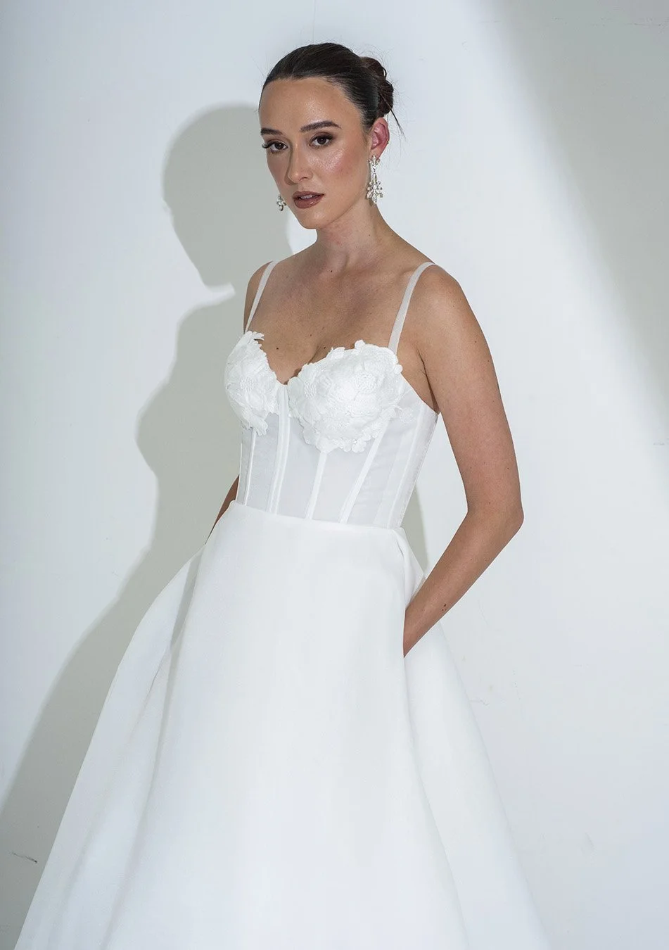 A woman wearing a white wedding dress with thin straps, floral lace details on the bodice, and a full skirt. She has dark hair styled in an updo and is wearing earrings, standing against a plain white wall.
