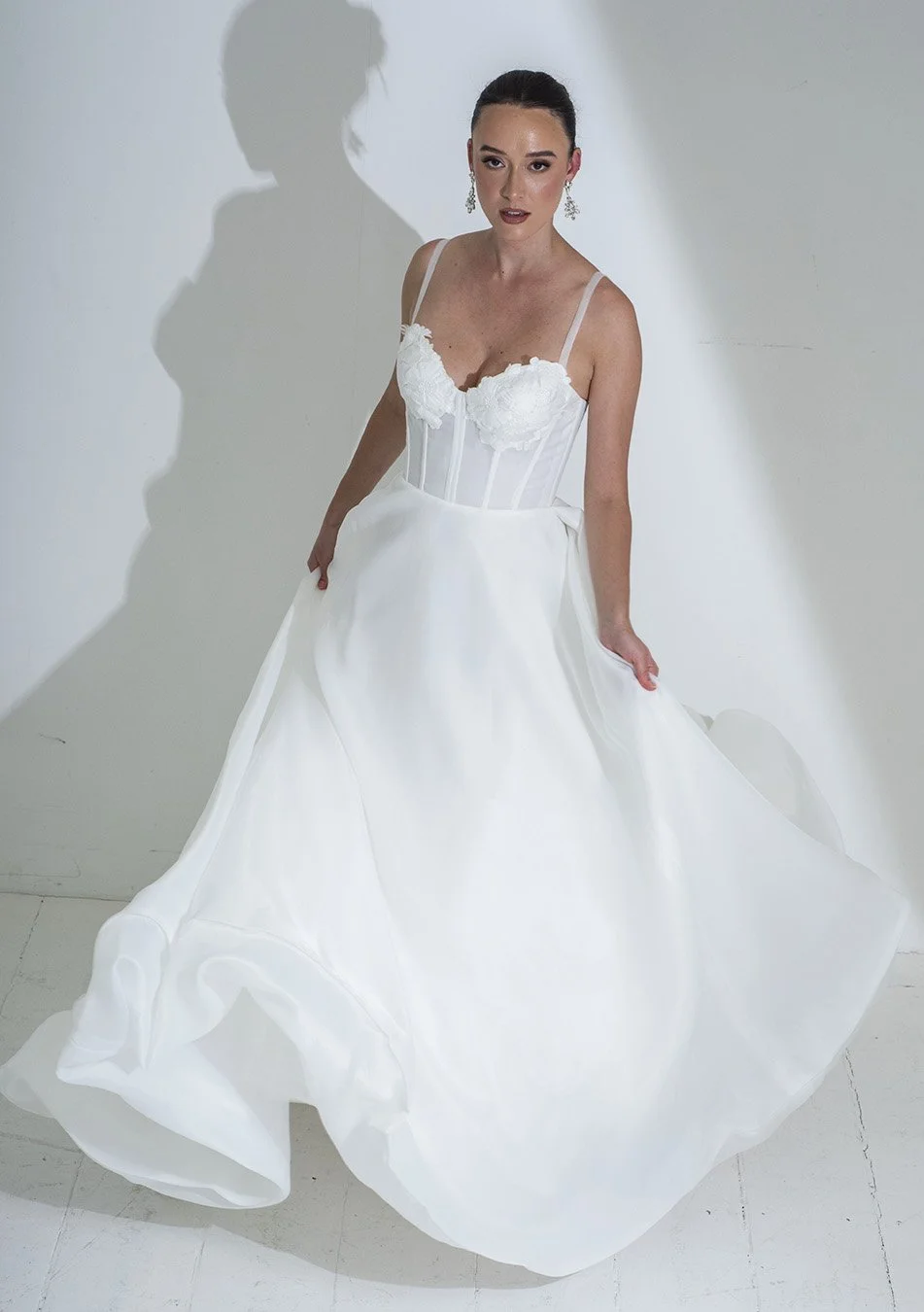A woman in a white wedding dress with lace details and spaghetti straps, standing against a plain white wall, casting a shadow on the wall.