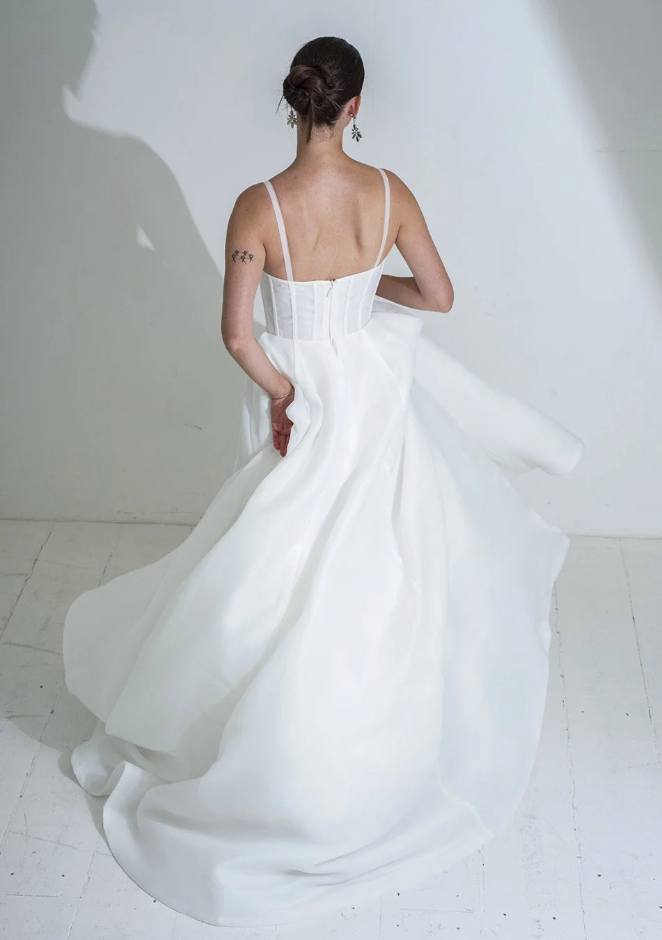 A woman wearing a white wedding dress with spaghetti straps, standing with her back to the camera, holding up part of her dress with her right hand, and standing against a plain white wall.