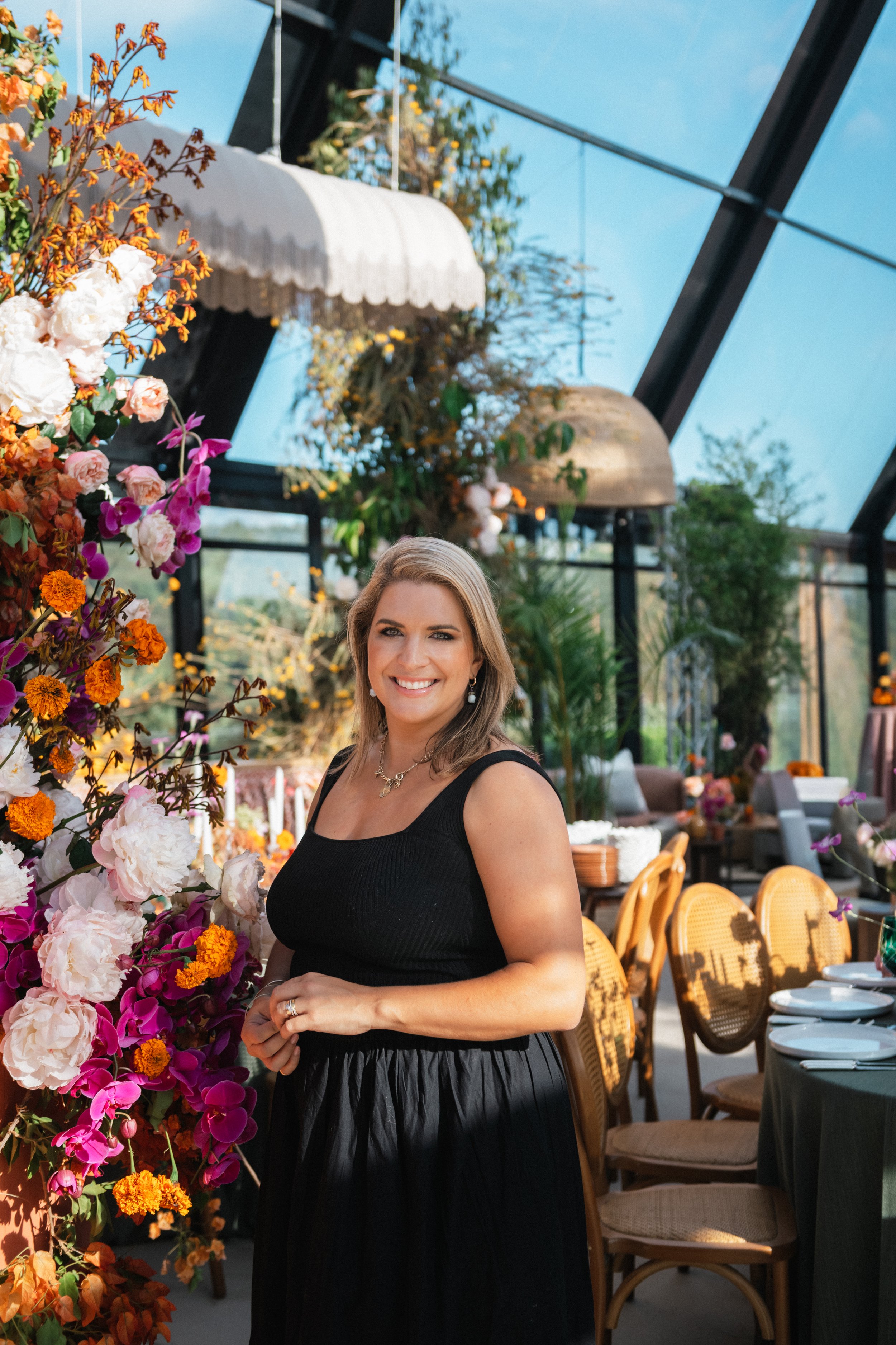 A woman smiling and standing in front of a floral display at a restaurant or event space with sunlight streaming in through large windows.