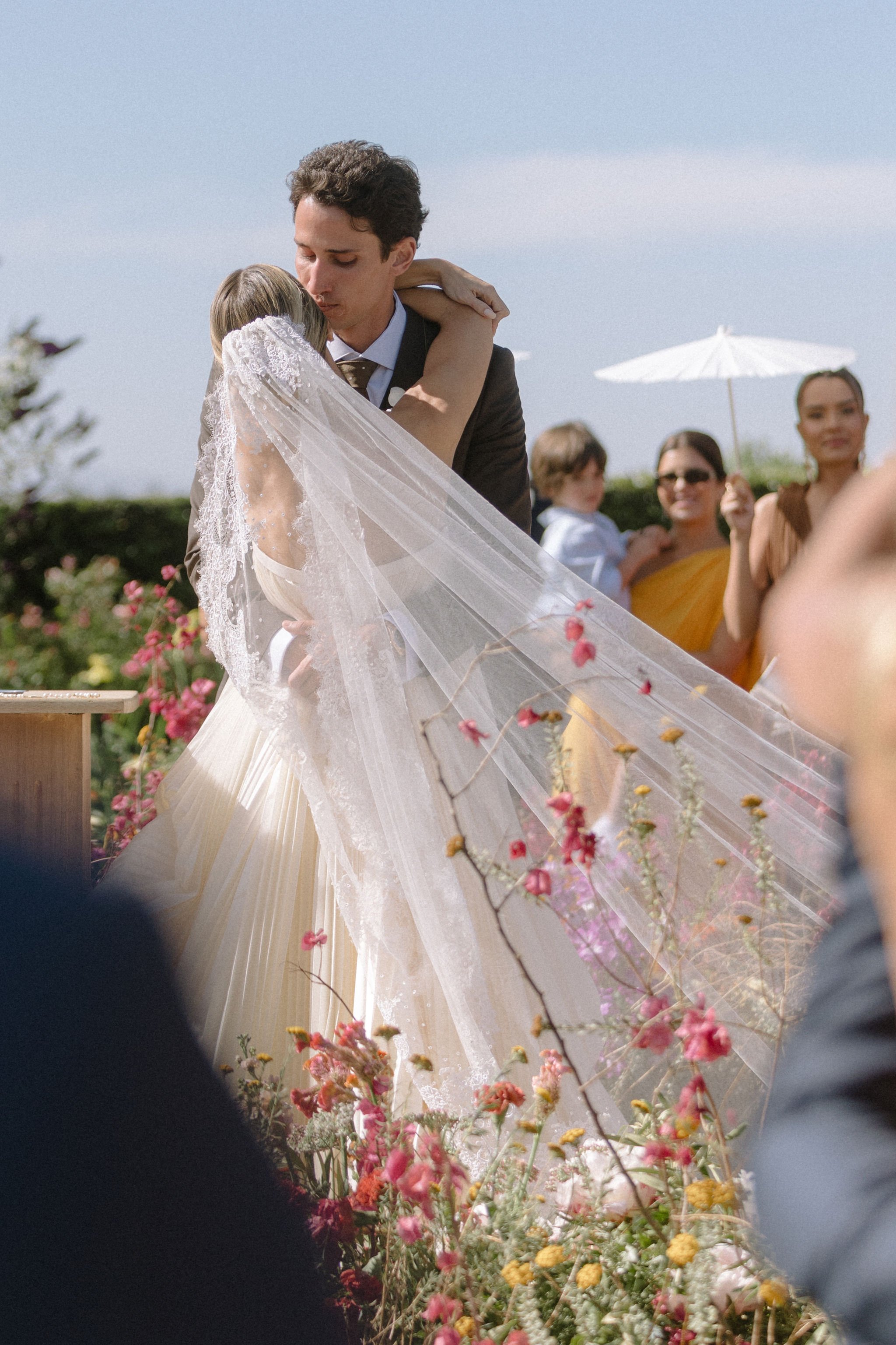 A couple is hugging during a wedding ceremony with colorful flowers and other guests in the background.