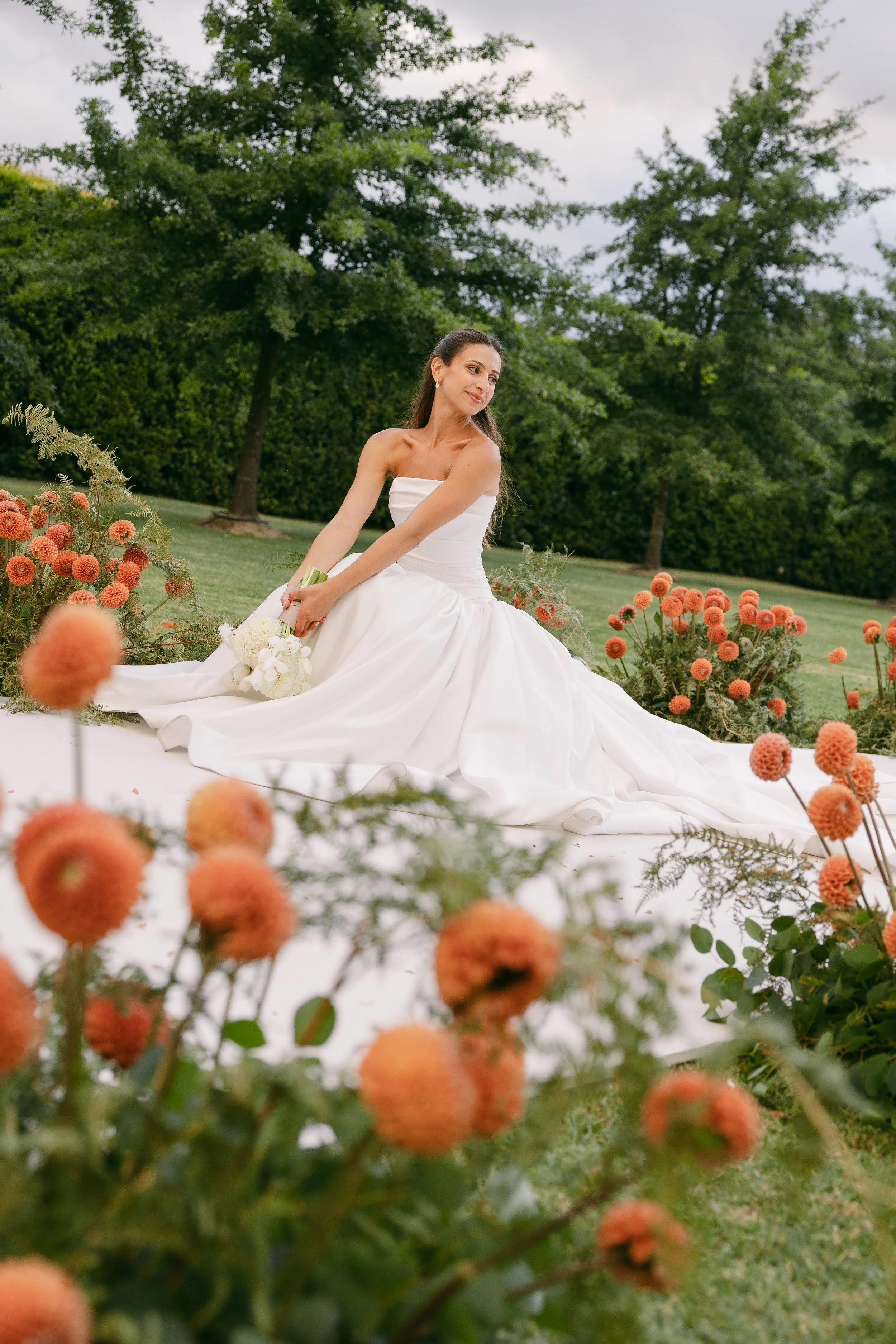 A bride in a white wedding dress sitting outdoors on a grassy area surrounded by orange pom-pom flowers. She is holding a small bouquet of white flowers and looking slightly to the side with a gentle smile. In the background, there are tall green trees and a cloudy sky.