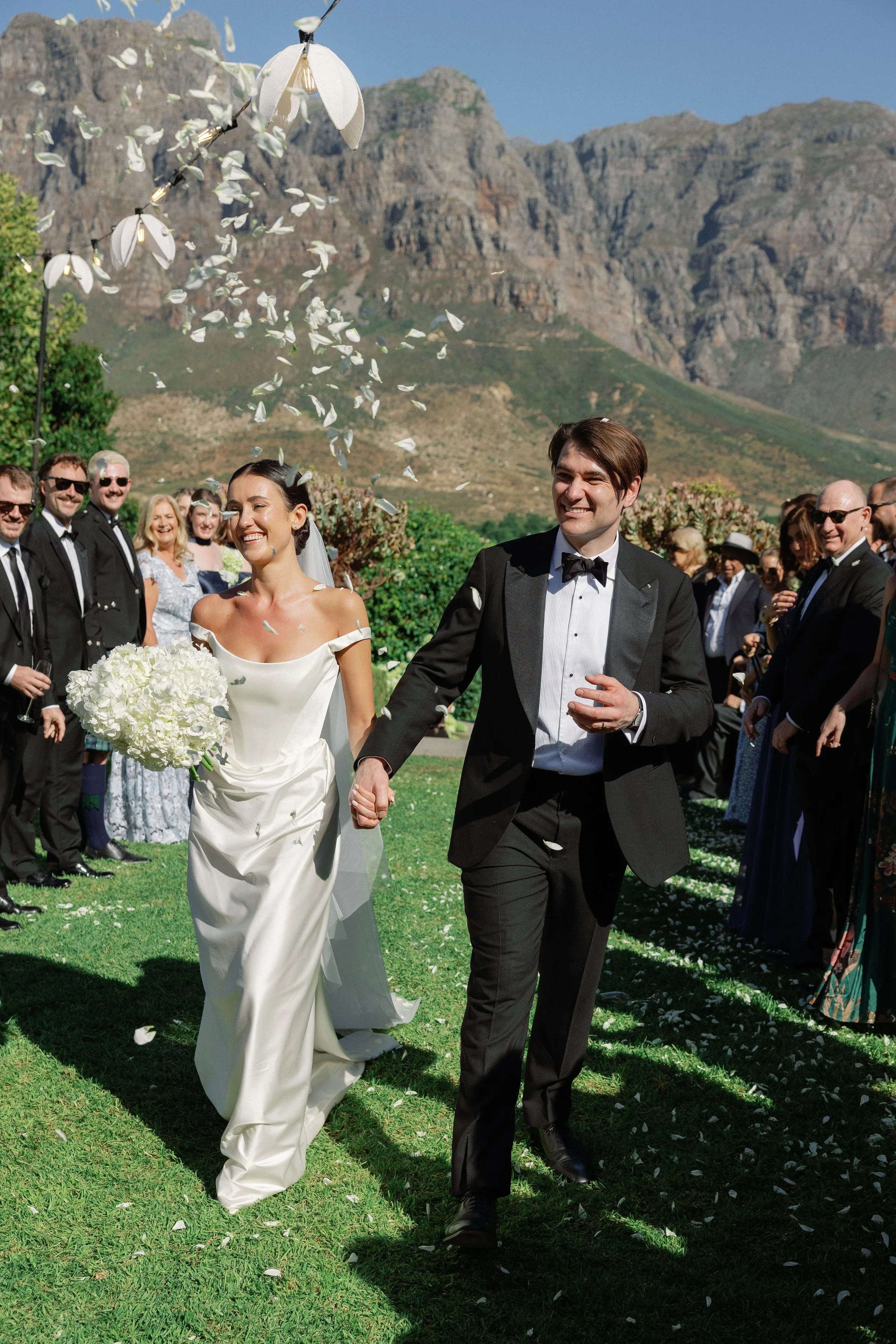 A bride and groom walking hand in hand outdoors during their wedding ceremony, smiling, with guests in the background and mountains in the distance, amid falling white flower petals.