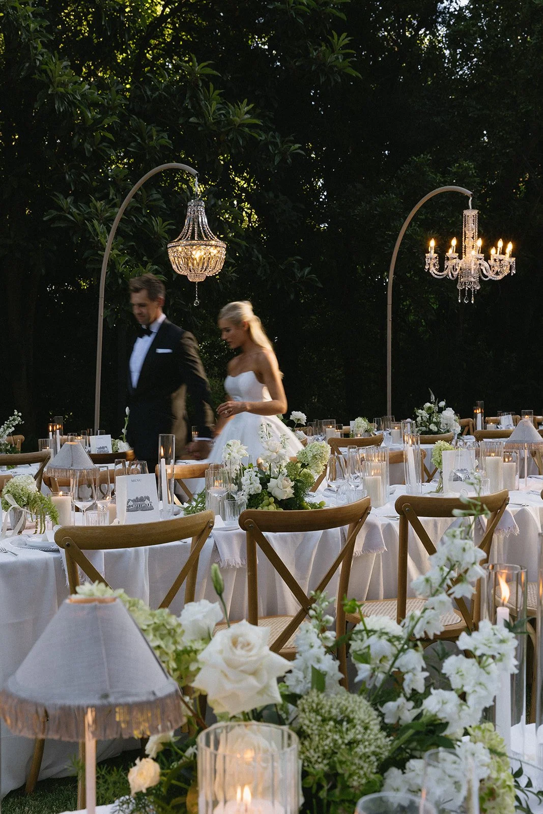 Elegant outdoor wedding reception with tables decorated with white flowers, candles, and table settings under large chandeliers in a lush green garden.