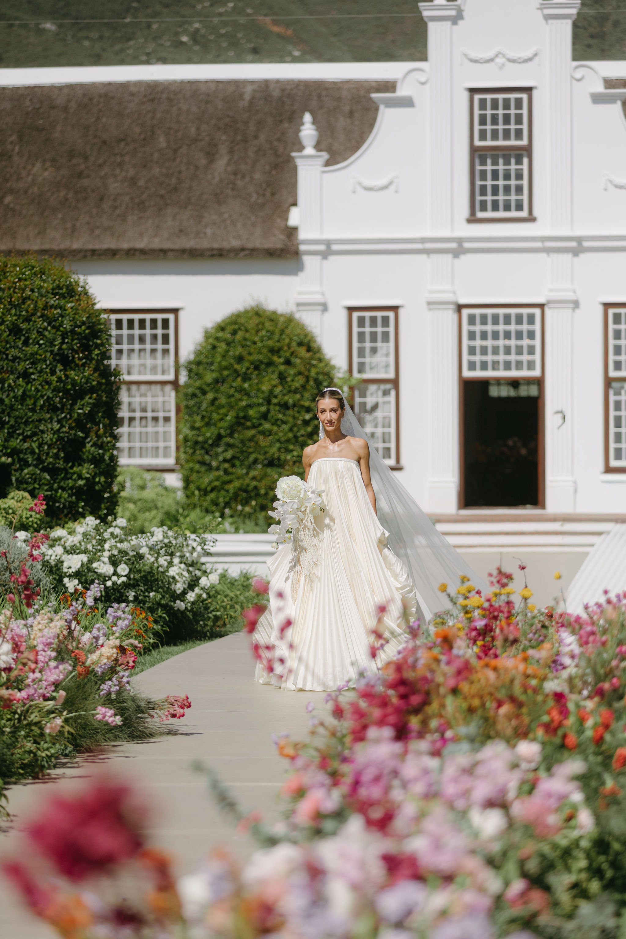 A bride in a strapless cream-colored wedding gown with pleated skirt walking on a pathway lined with colorful flowers in front of a white building with large windows and a brown thatched roof.