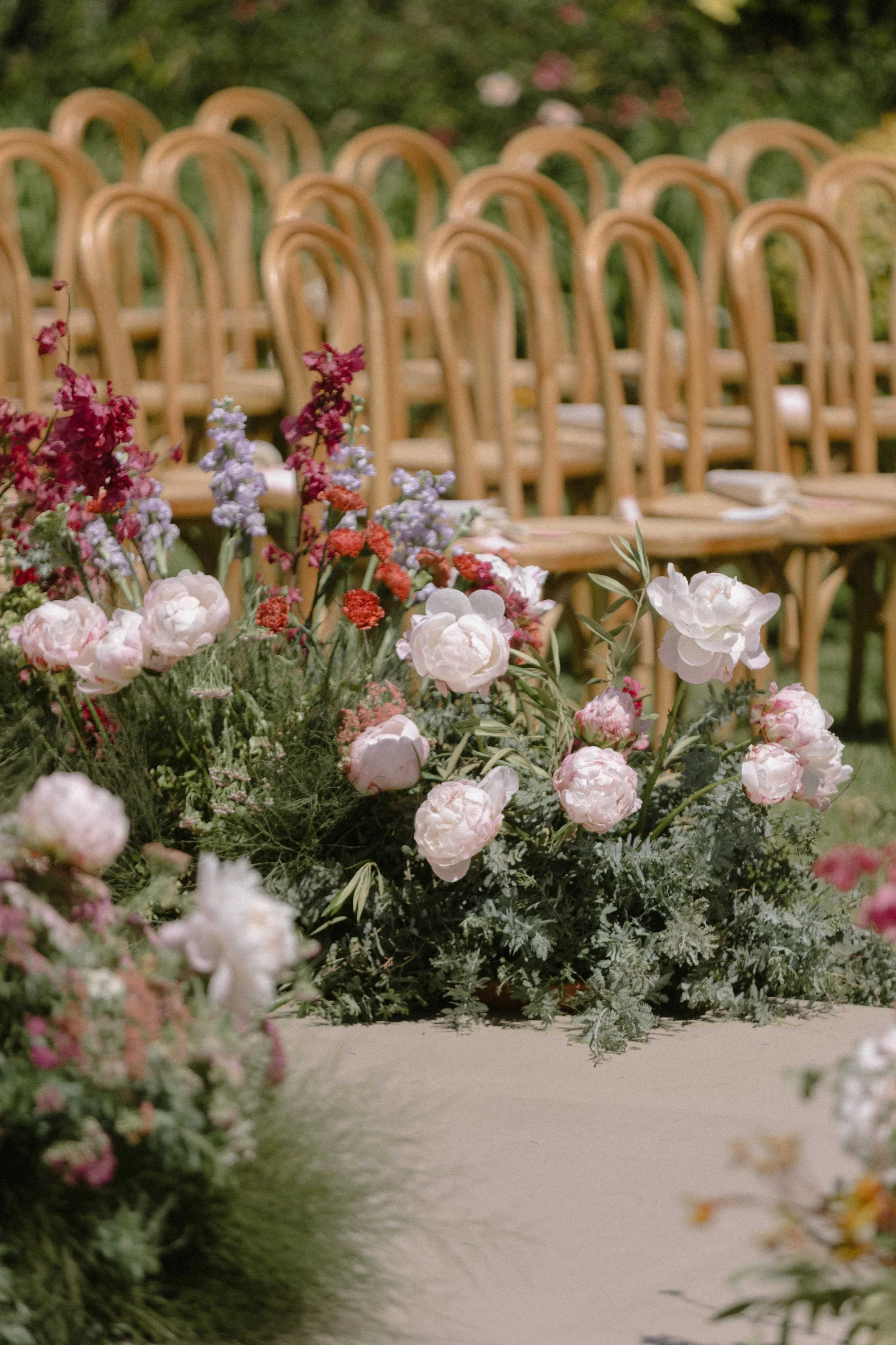 Rows of empty wooden chairs with floral arrangements on the aisle in a garden or outdoor setting.