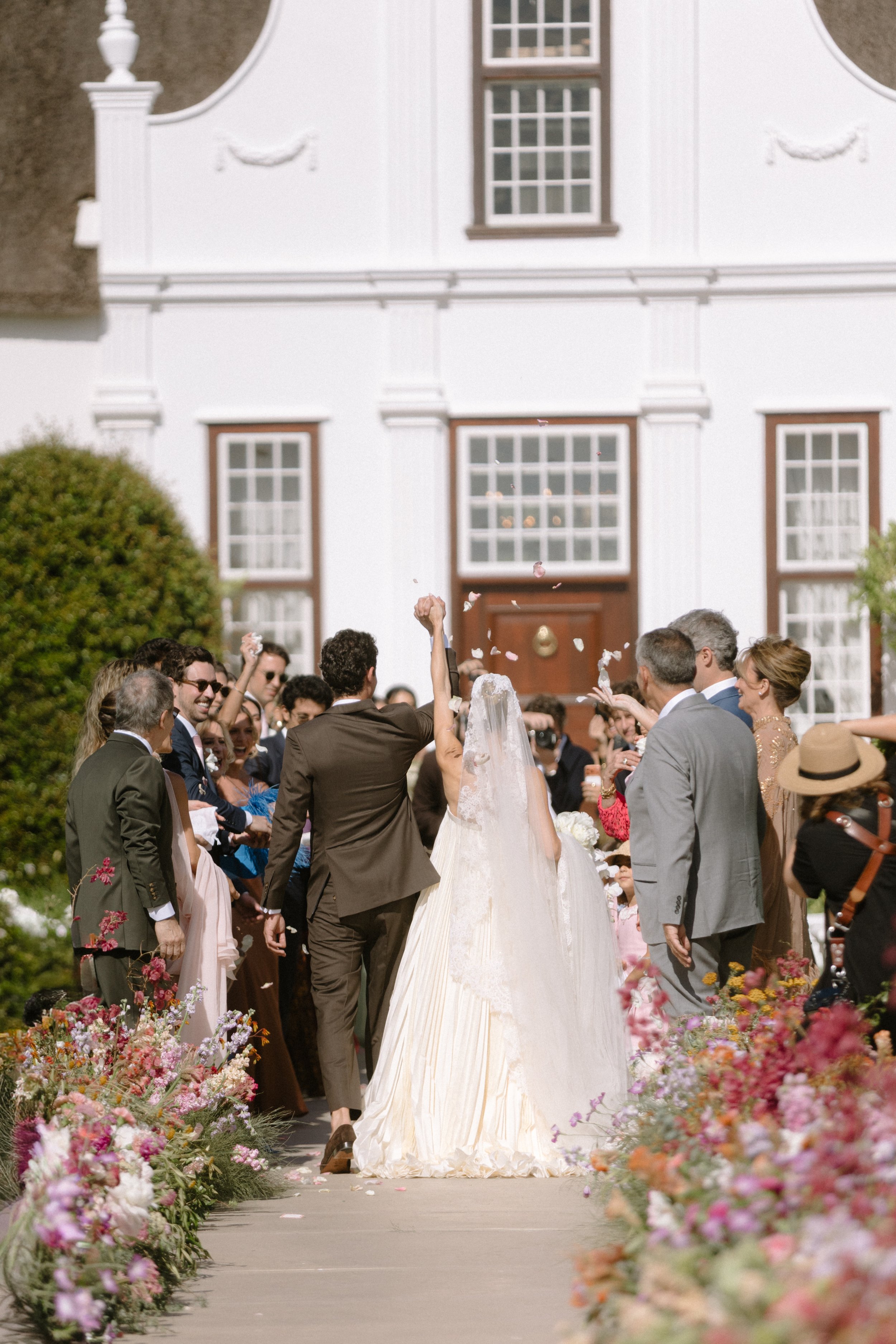 A wedding celebration outside a large white house with a group of people surrounding the bride and groom. The bride is in a white dress, and the groom is in a brown suit. Flower arrangements line the walkway, and people are throwing flower petals into the air.