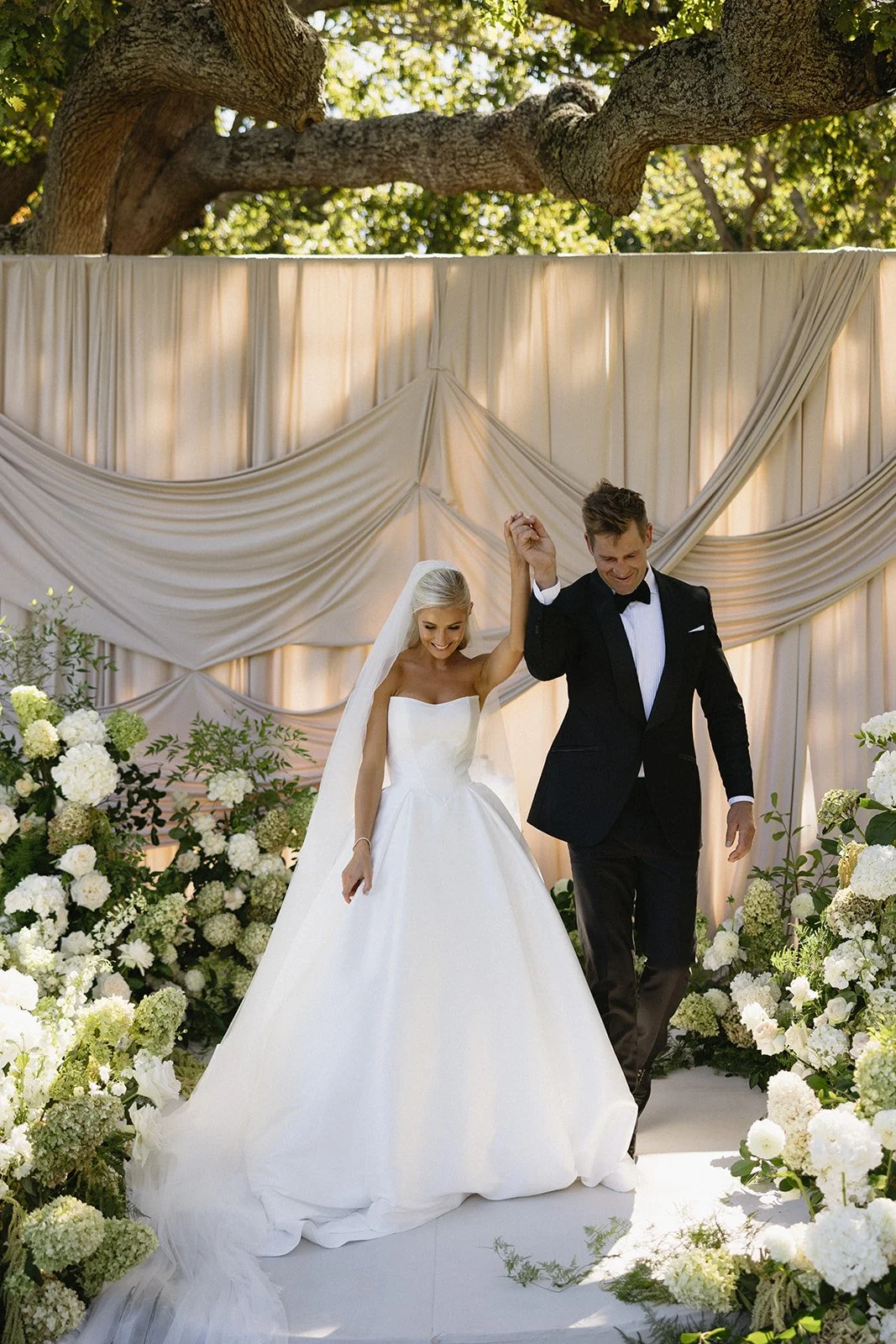 A bride and groom walking hand in hand down the aisle at an outdoor wedding, surrounded by white flowers and greenery, with a draped fabric backdrop and a large tree overhead.