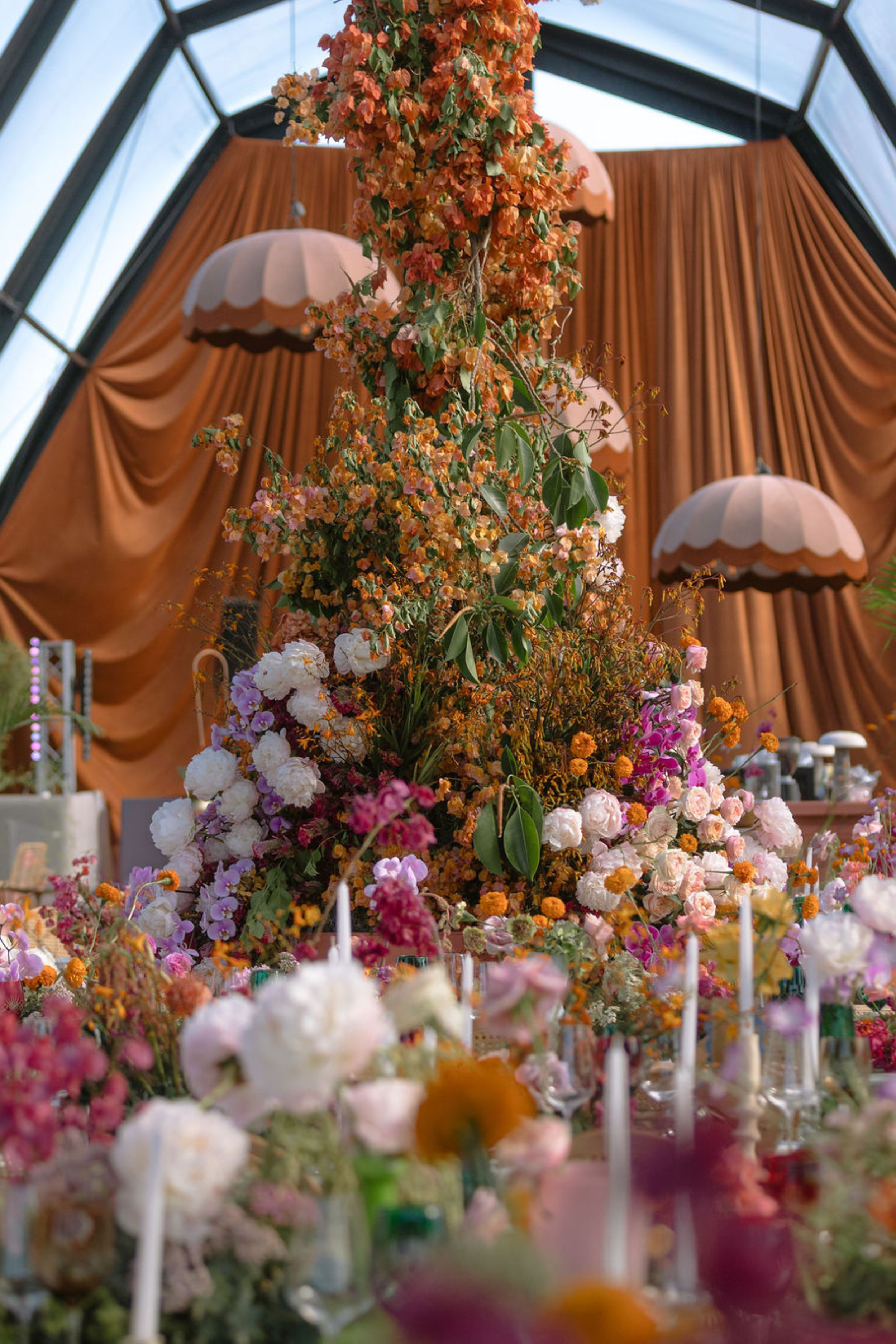 A tall floral arrangement with pink, orange, and white flowers and green leaves, set against a backdrop of orange curtains and vintage-style hanging lamps inside a glass conservatory.