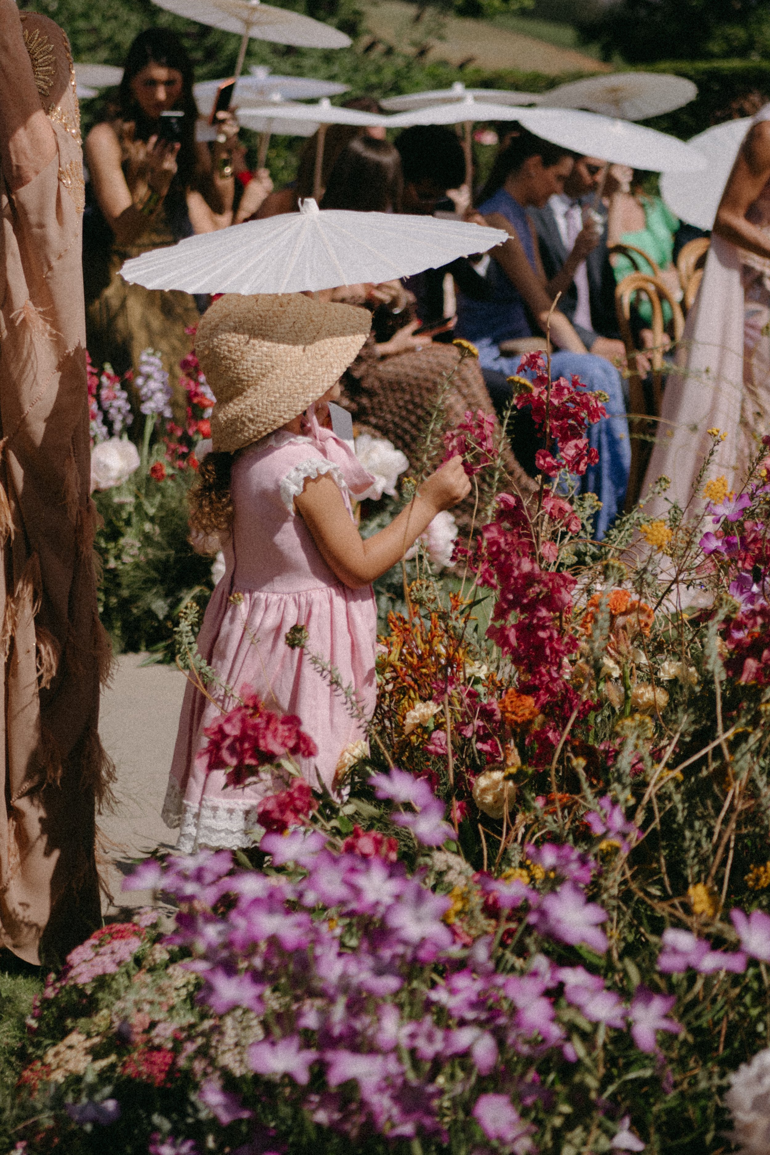 People sitting on chairs outdoors at a garden or flower show, with white parasols overhead and colorful flowers in the foreground.