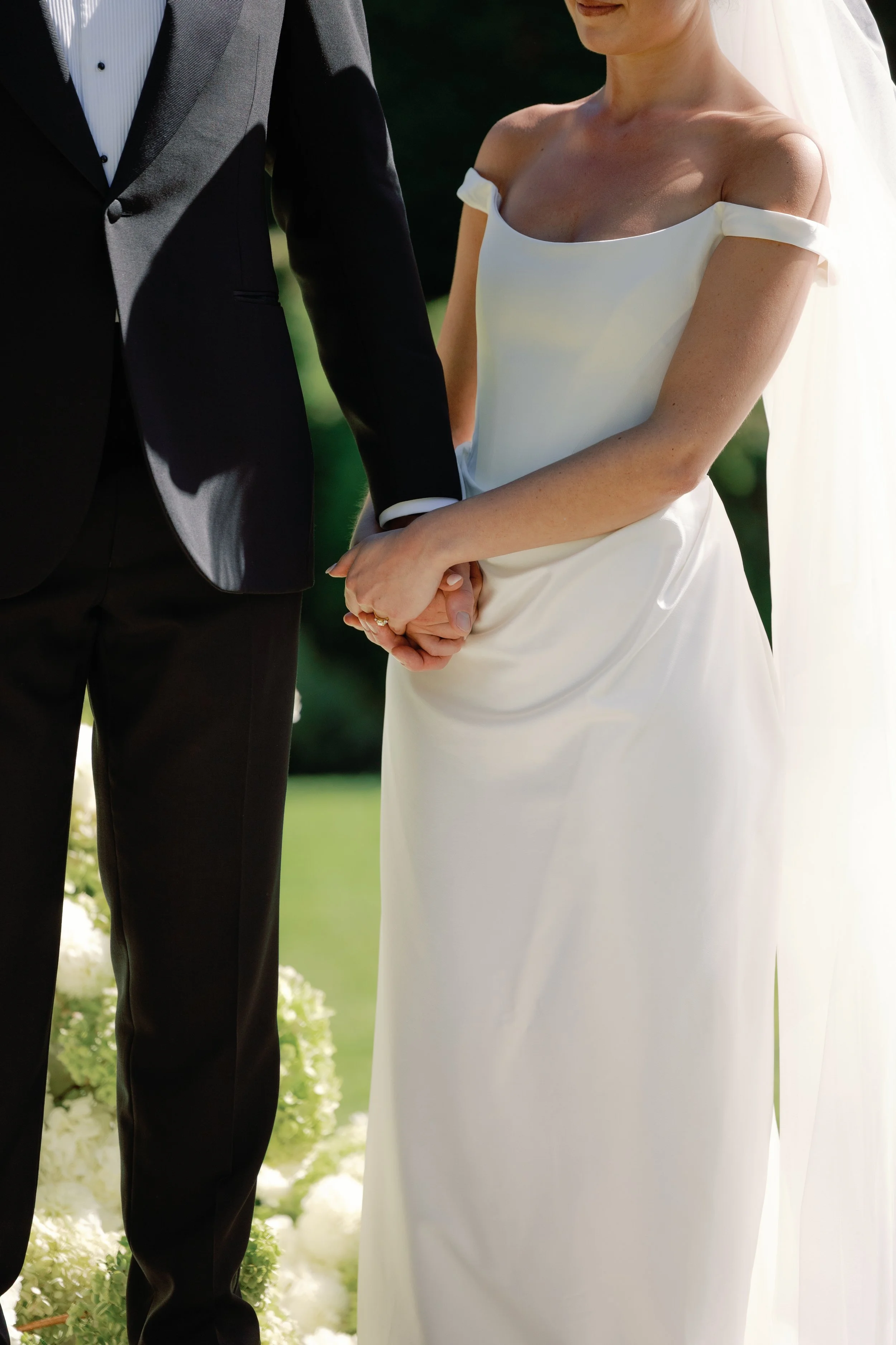 Bride and groom holding hands during wedding ceremony, bride in white dress with off-the-shoulder sleeves and veil, groom in black tuxedo with white shirt.