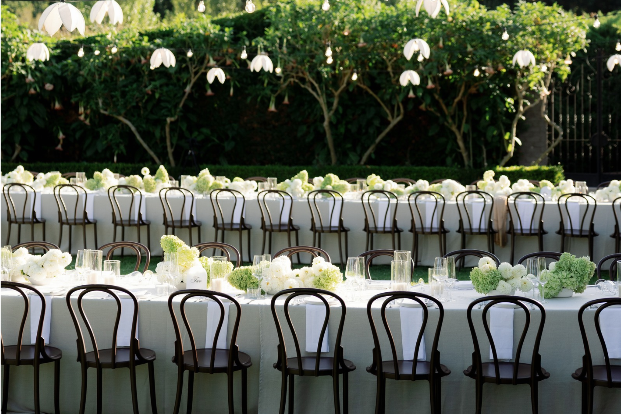 Long rectangular banquet table with white tablecloth and floral centerpieces of white and green flowers, set with glasses and cutlery, outdoor setting with green bushes and string lights overhead, surrounded by black chairs.