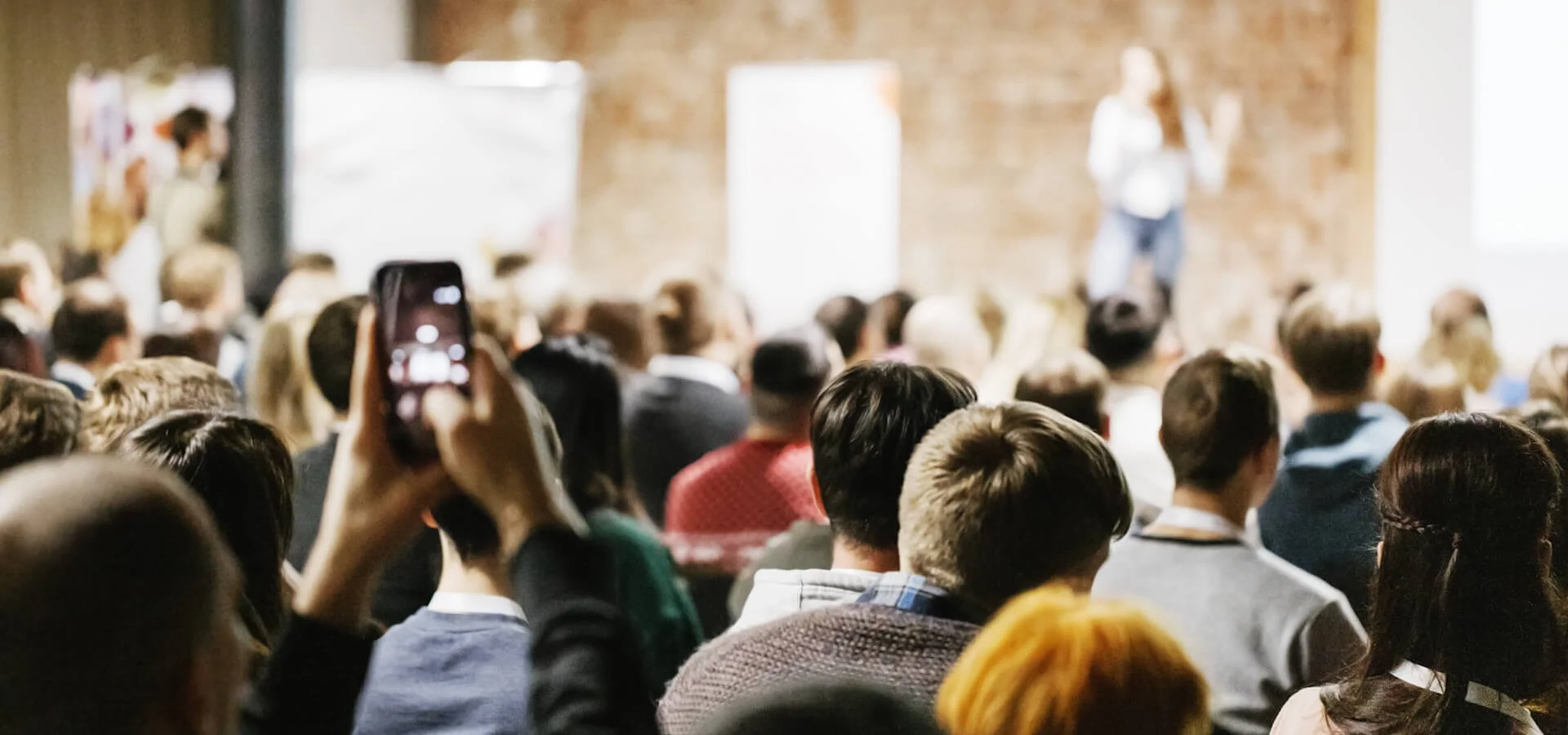 Crowded audience at a live event with speaker on stage near projector screen, one attendee filming with phone, highlighting event videography challenges