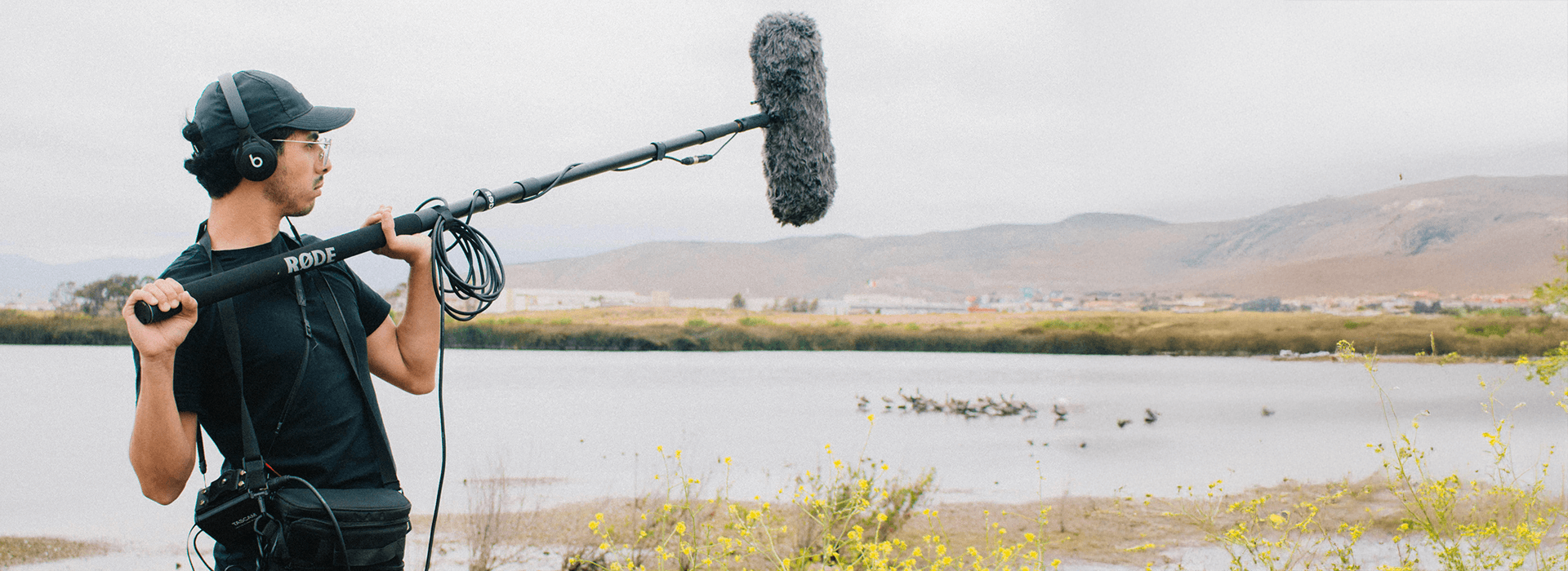 A sound recordist wearing a black cap, headphones, and black shirt stands outdoors holding a long boom pole with a large fluffy windscreen (dead cat) microphone, pointing it toward a scenic river with mountains in the background.