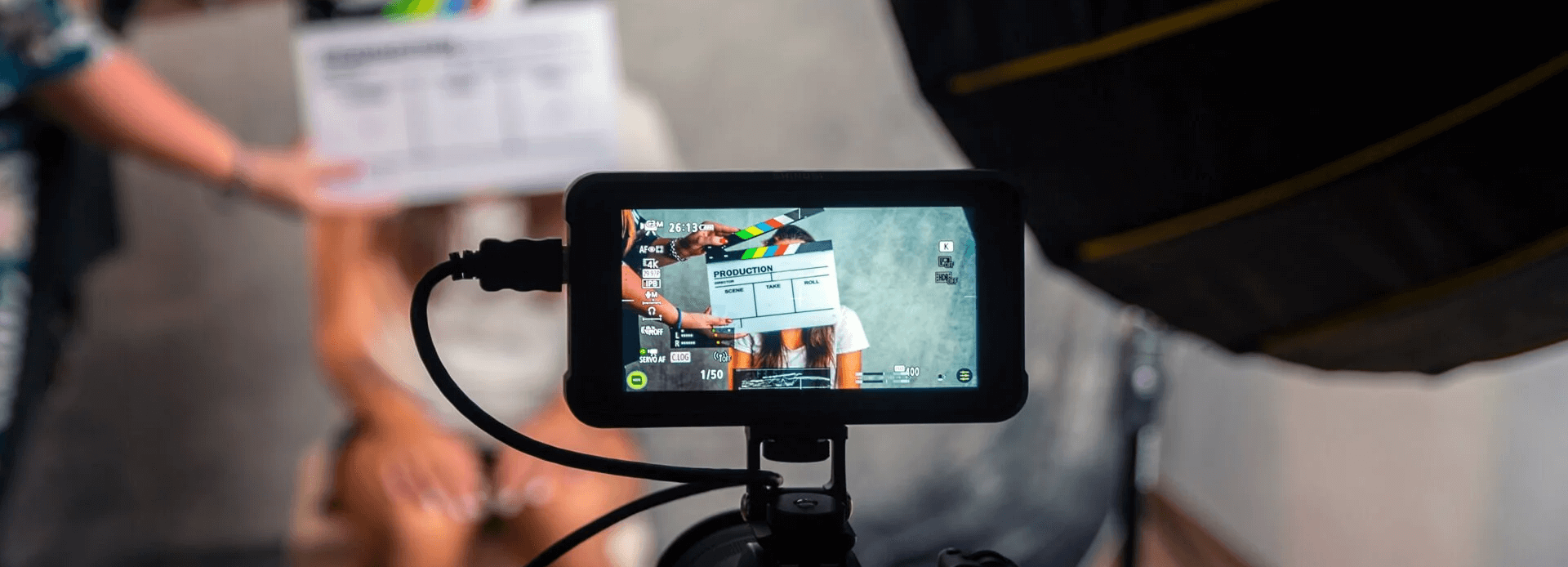 Close-up of a camera monitor displaying a clapperboard and subject during filming, with crew members holding a script and slate in the background. Illustrates the active shooting process in the production stage of video creation.