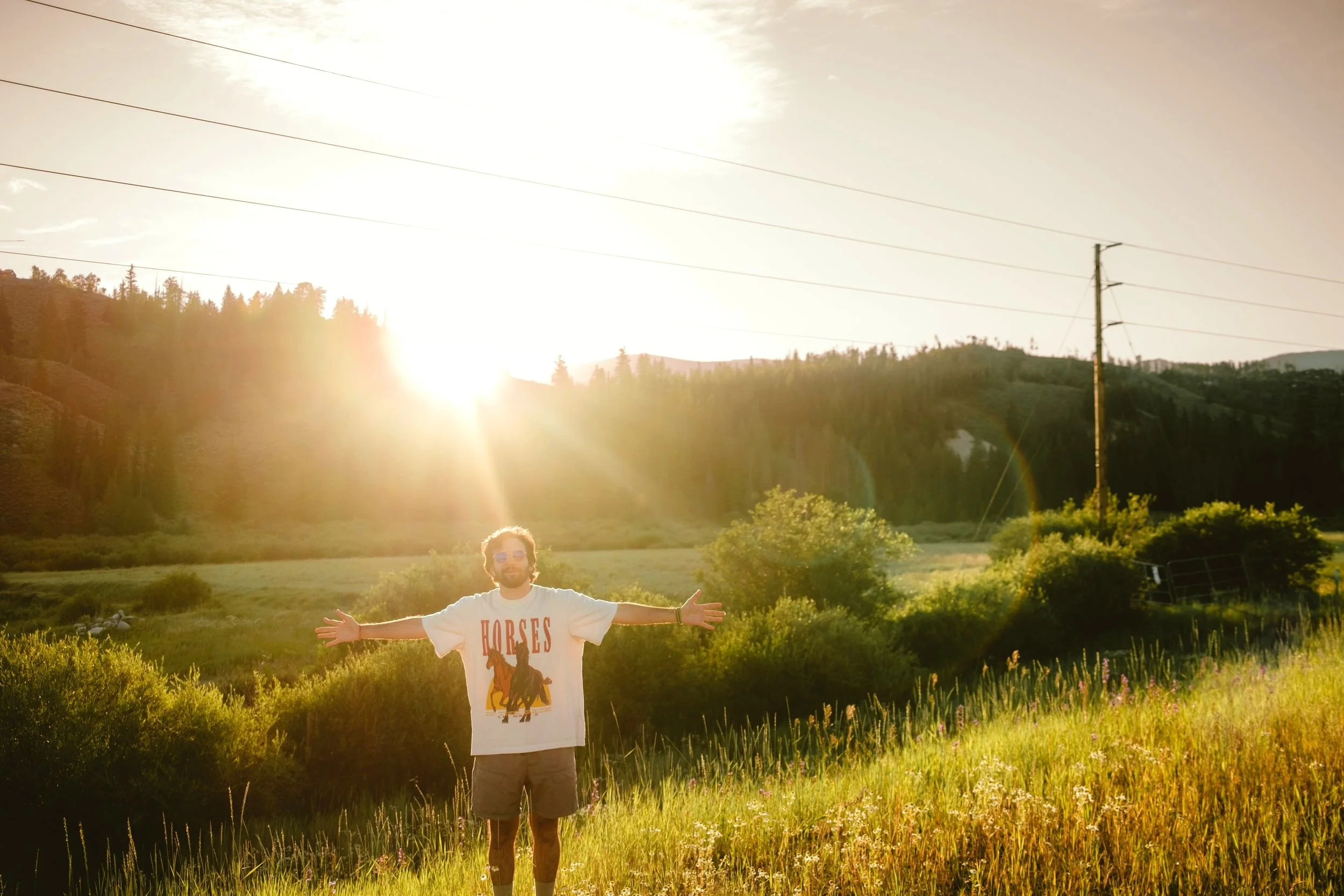 A man standing in a field with his arms outstretched during sunset, wearing sunglasses and a white T-shirt with a horse graphic and the word "HORSES" on it.