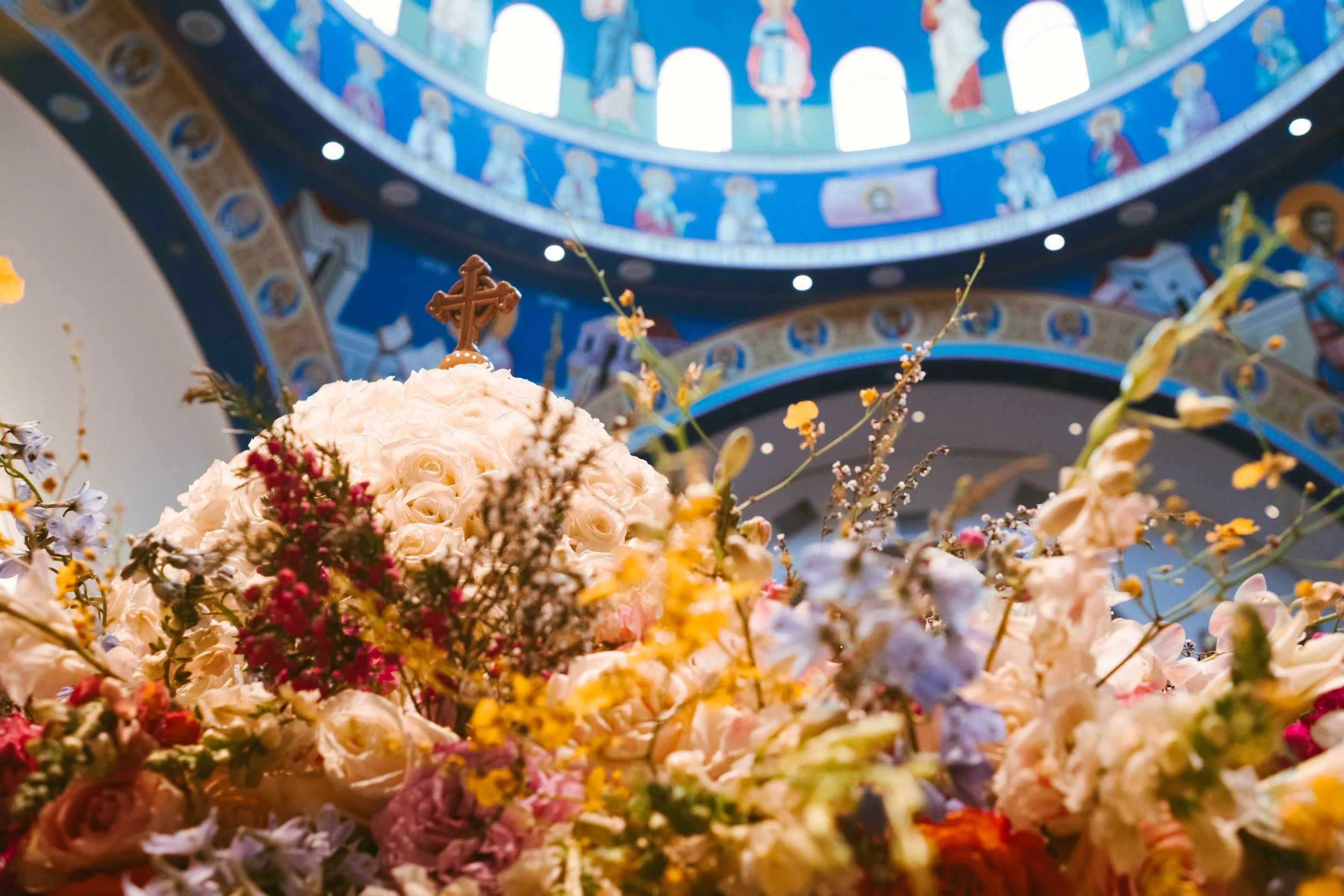 A floral arrangement with white roses, pink, purple, yellow, and cream-colored flowers, topped with a brown cross, inside a church with a blue dome and religious murals.