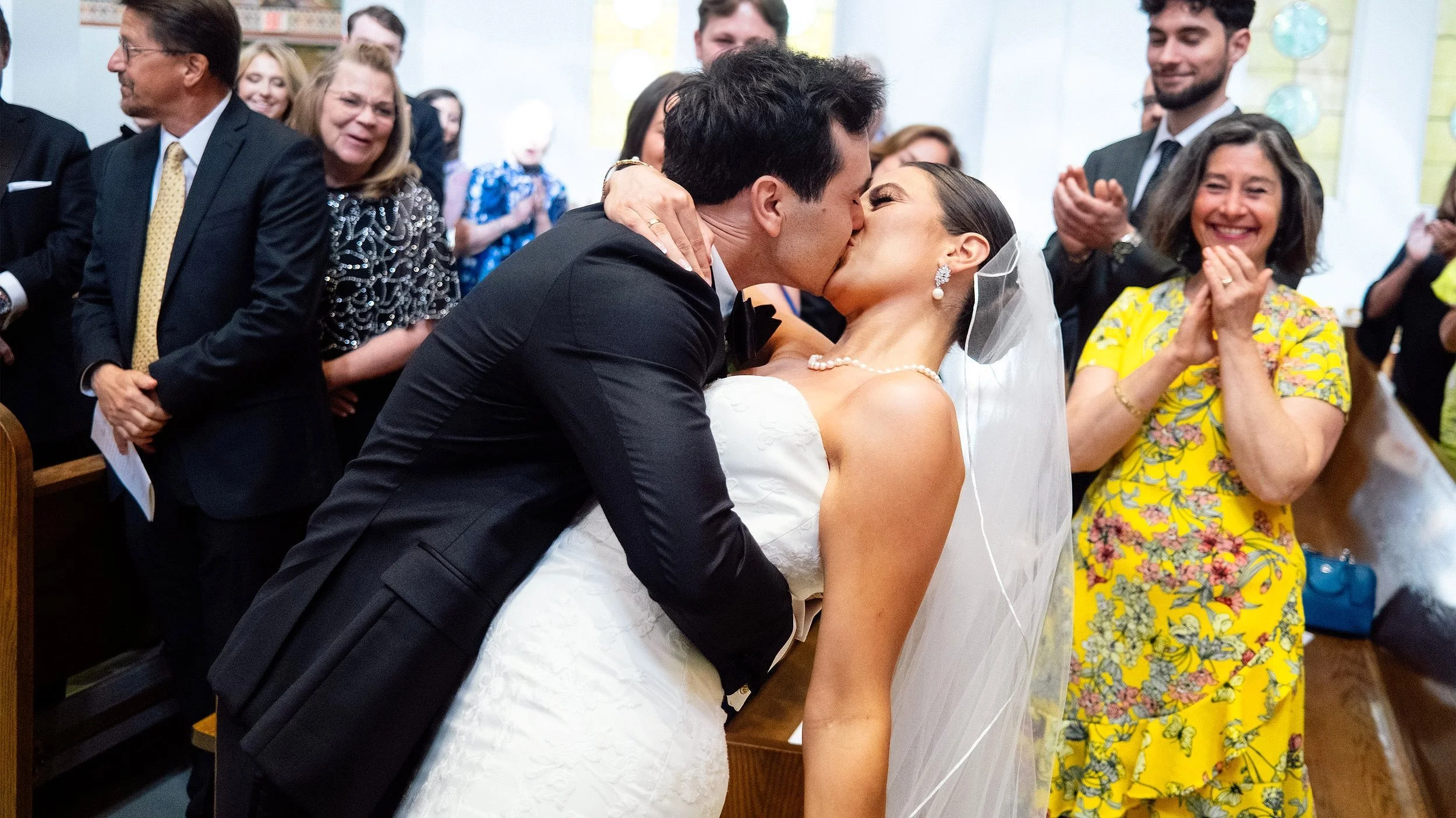 A bride and groom kissing during their wedding ceremony, surrounded by joyful guests in a church setting.