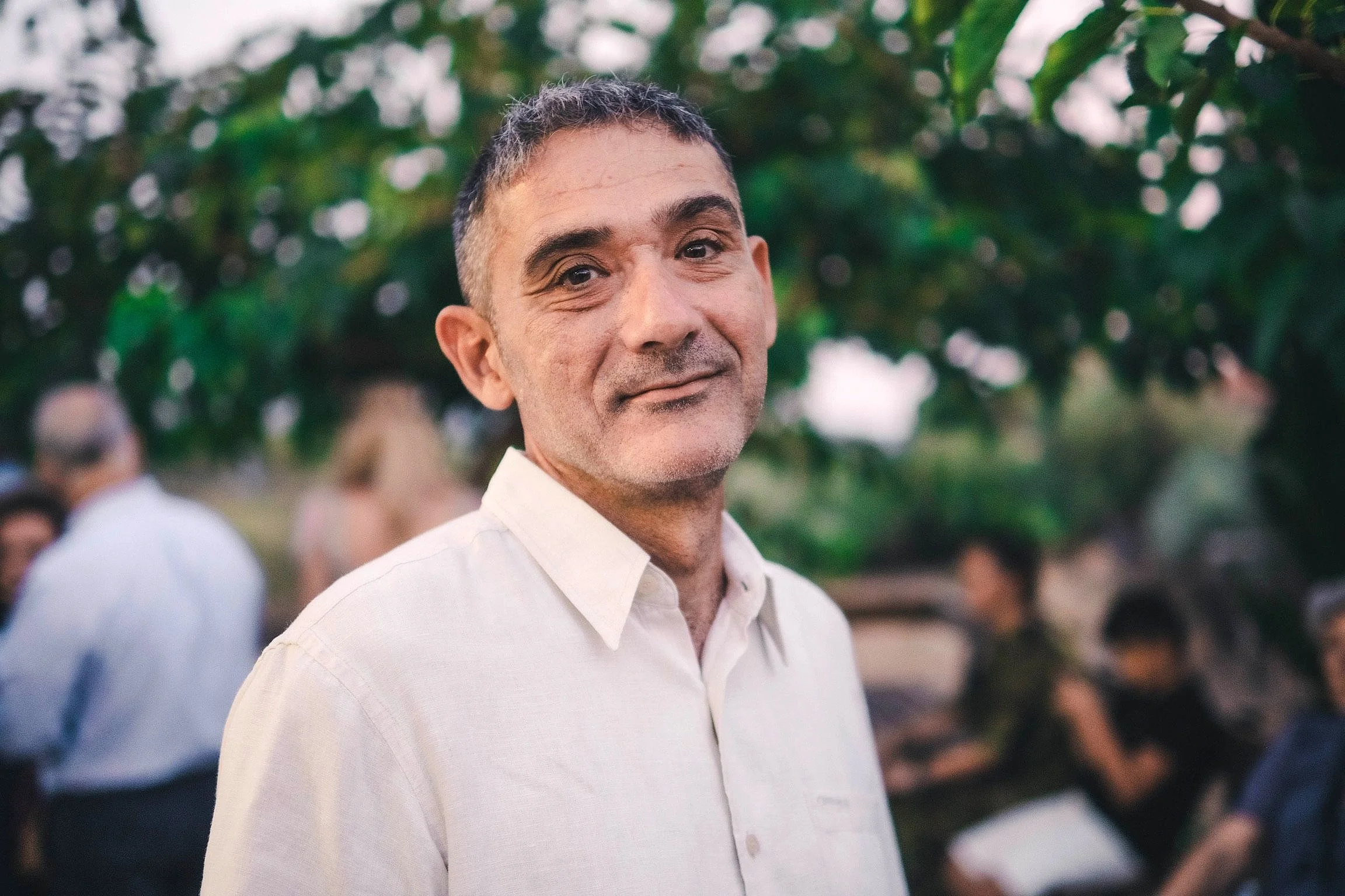 A middle-aged man with short dark hair and a slight smile, wearing a light-colored button-up shirt, standing outdoors with blurred people and greenery in the background.