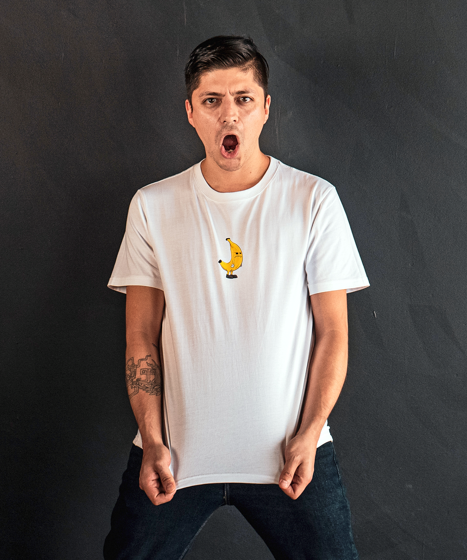 A young man with dark hair wearing a white T-shirt with a banana graphic, expressing surprise or anger, against a dark background.