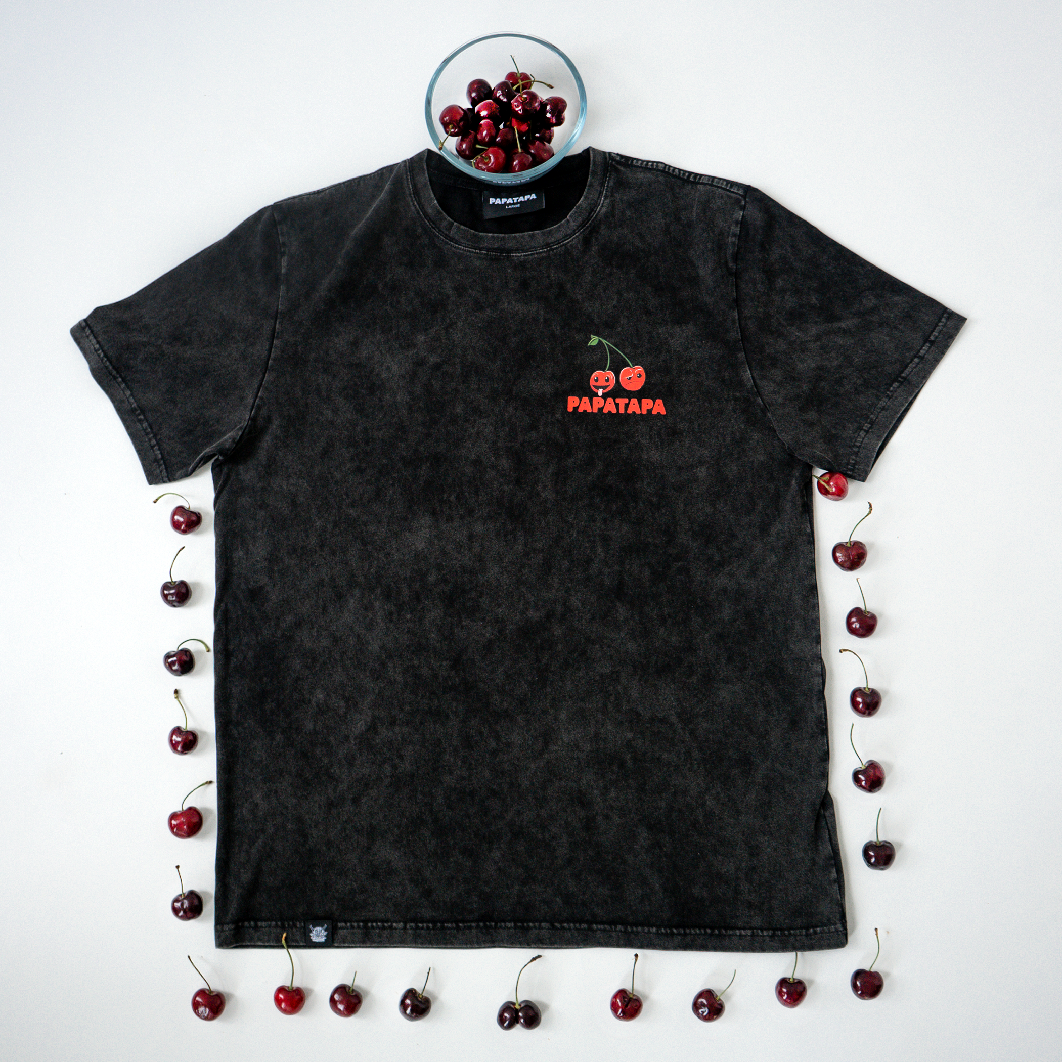 Black T-shirt with cherry graphic and text, surrounded by cherries, on a white background with a bowl of cherries placed on top.