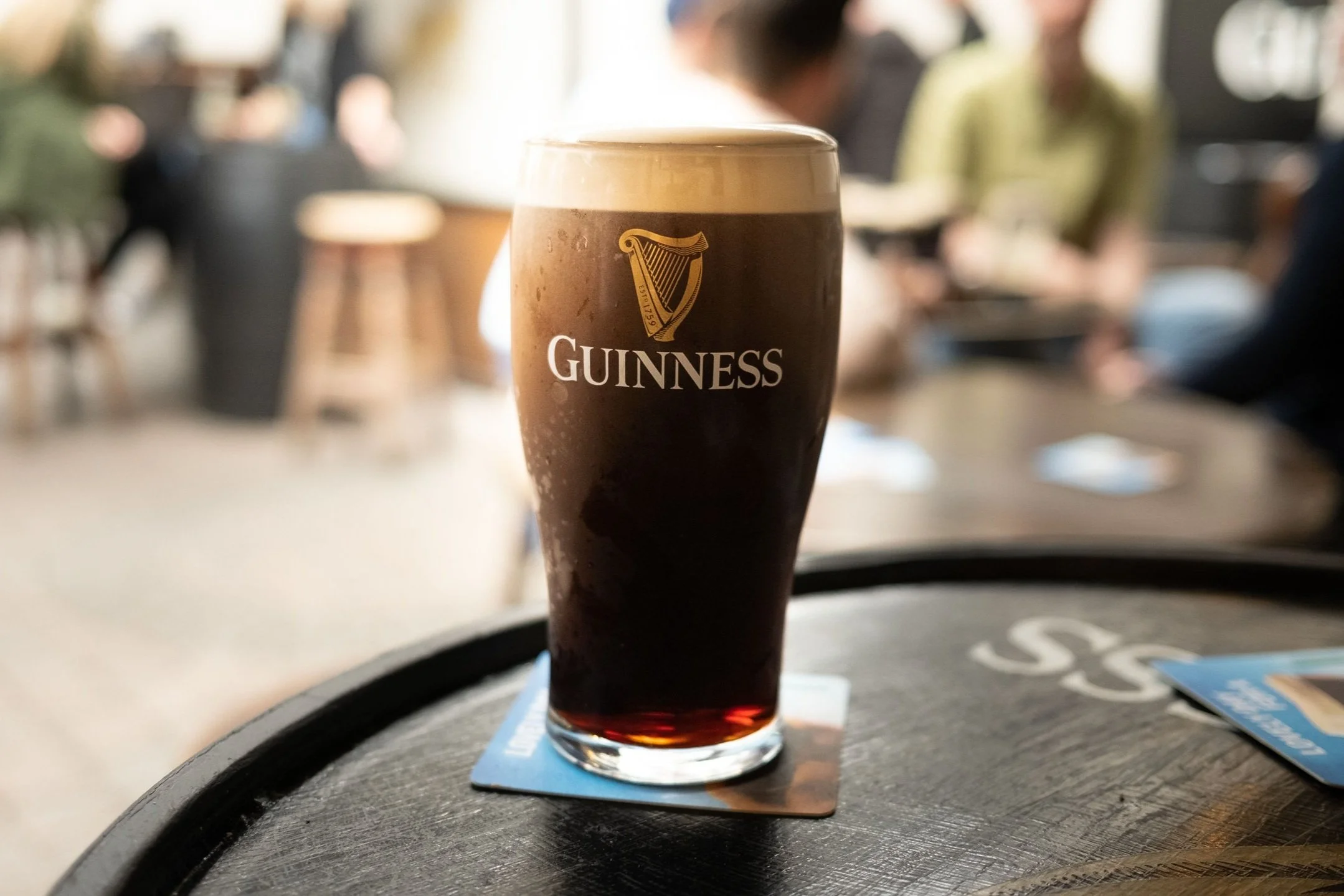 A pint glass of Guinness stout beer with a creamy head on a black round table, with a blurred background of people at a bar or pub.