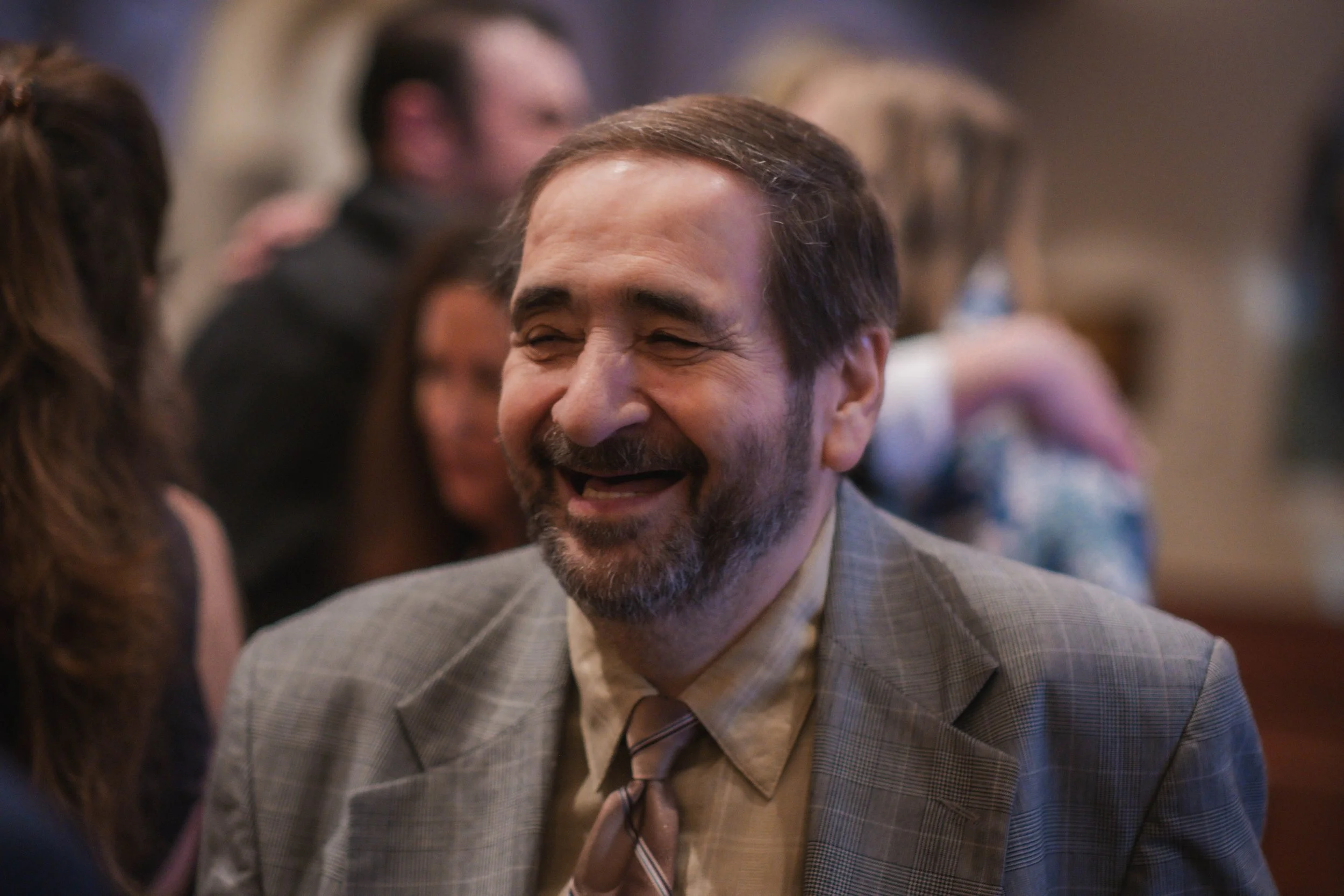 A middle-aged man with a beard and wearing a plaid suit and tie, smiling and laughing at a social gathering or party, surrounded by other people.